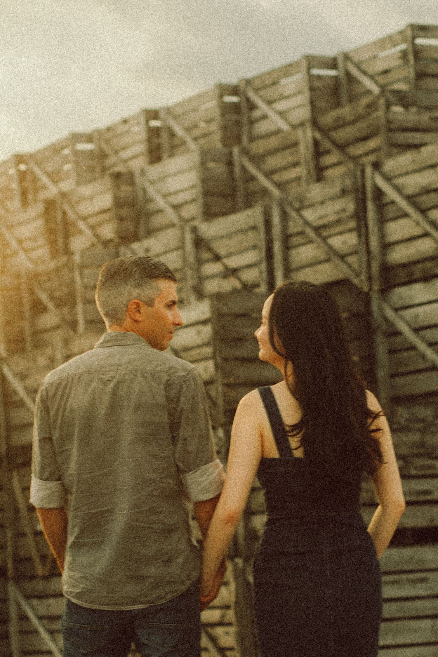 A couple holds hands while standing before a rustic wooden structure in warm sunset light.