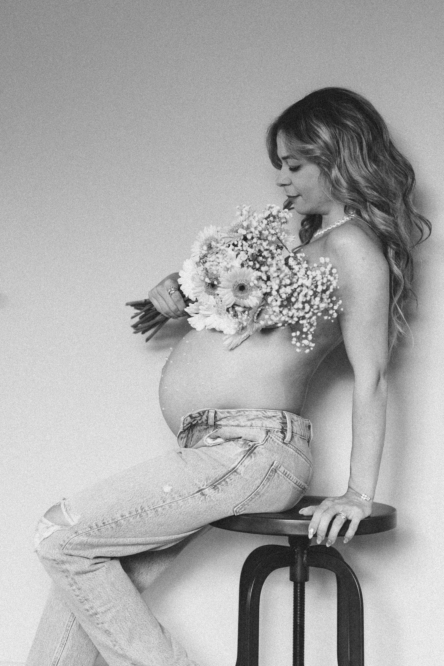Black and white maternity photo of pregnant woman holding flowers, wearing jeans, seated on stool.