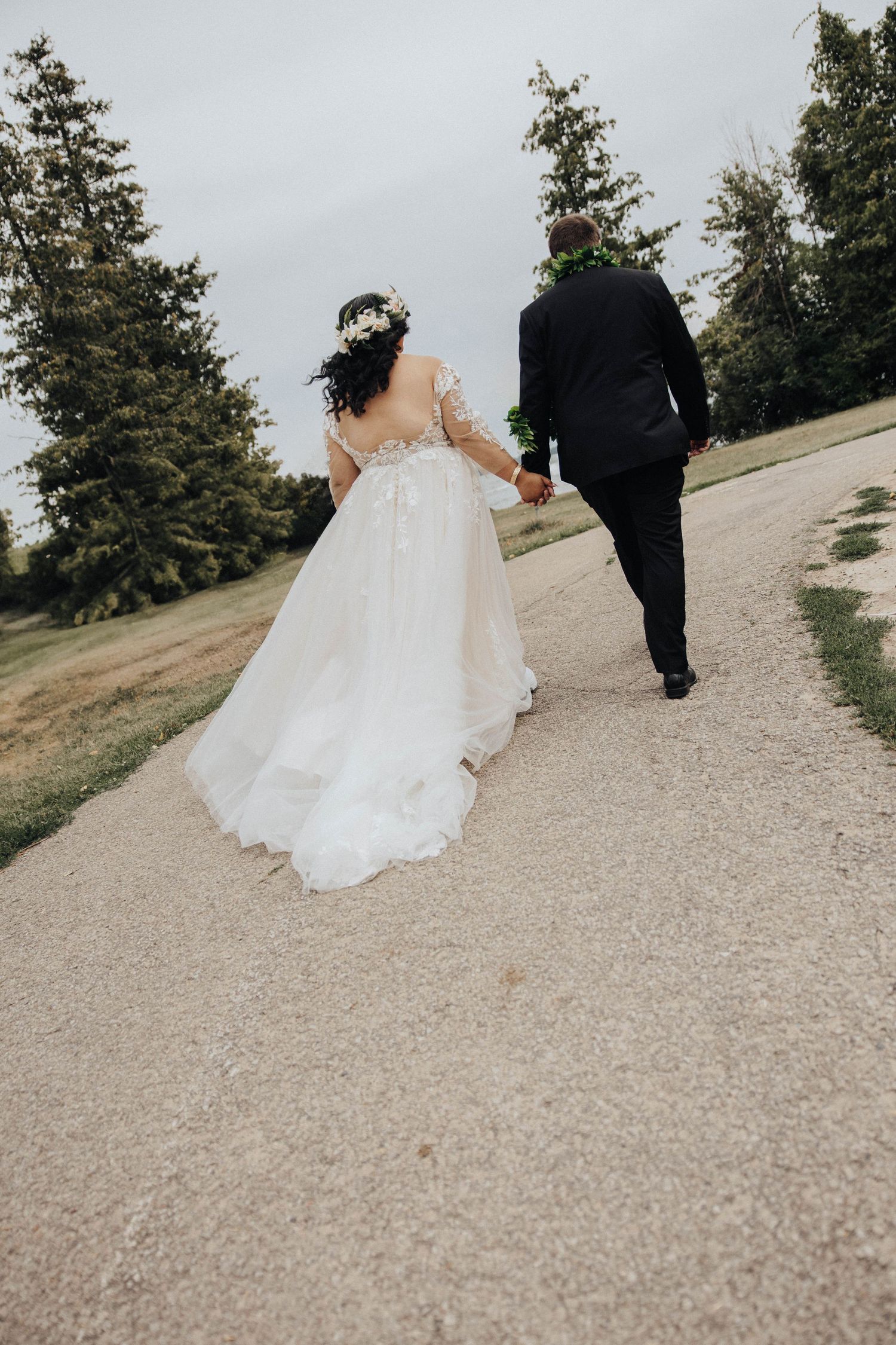 Newlywed couple walks together down a winding gravel path surrounded by evergreen trees on their wedding day.