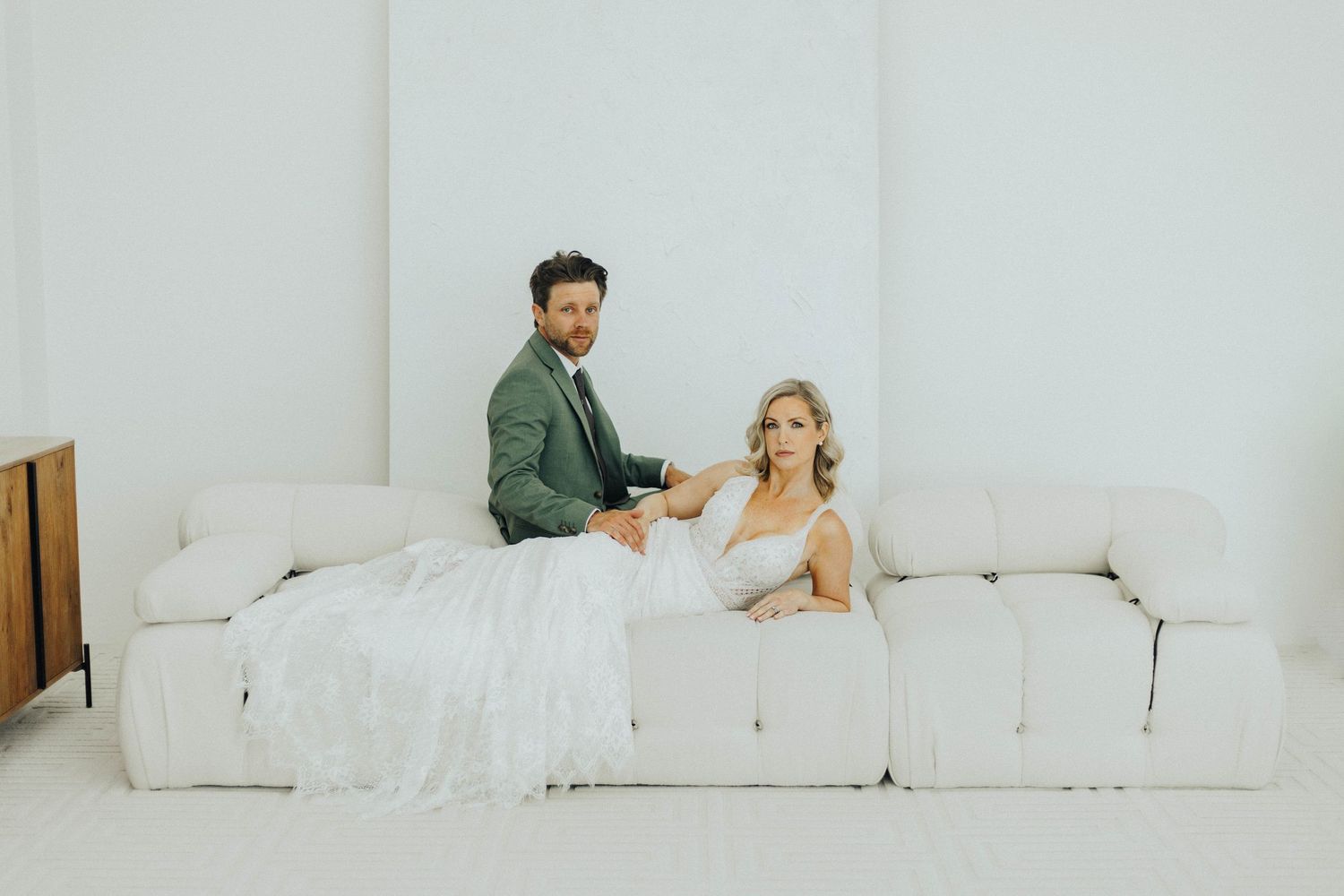 Professional wedding photography of couple relaxing on white couch during romantic indoor photo session.