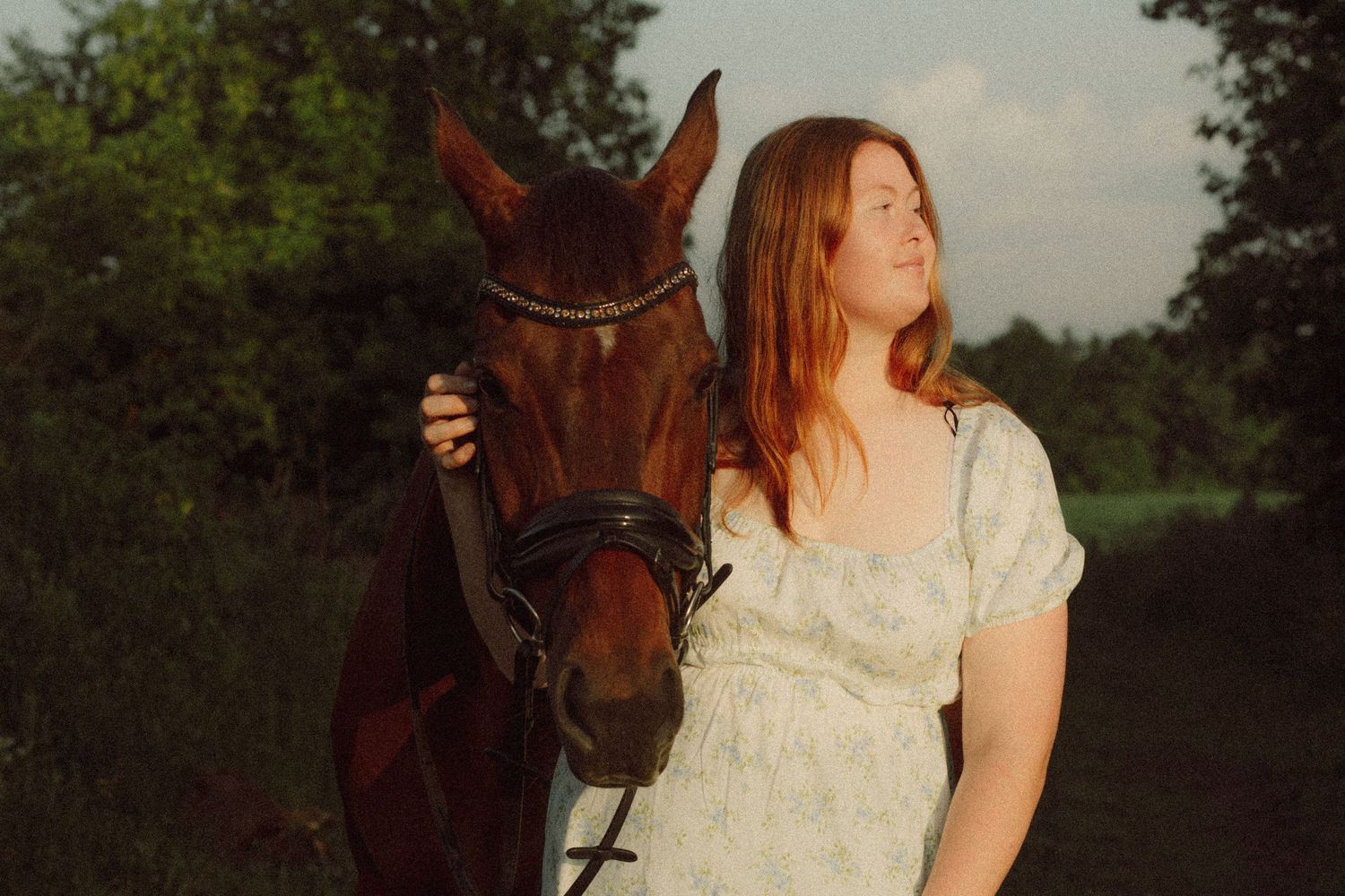 Young woman in white dress standing beside a chestnut horse outdoors at golden hour.