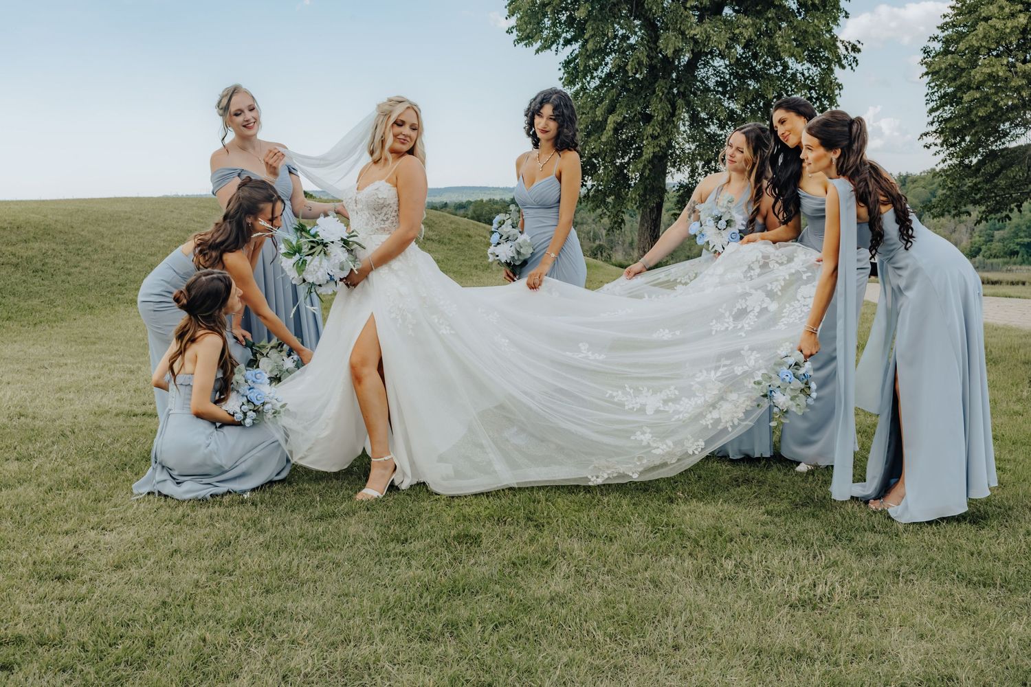 Bride in white gown surrounded by bridesmaids in blue dresses holding her train on a green lawn.