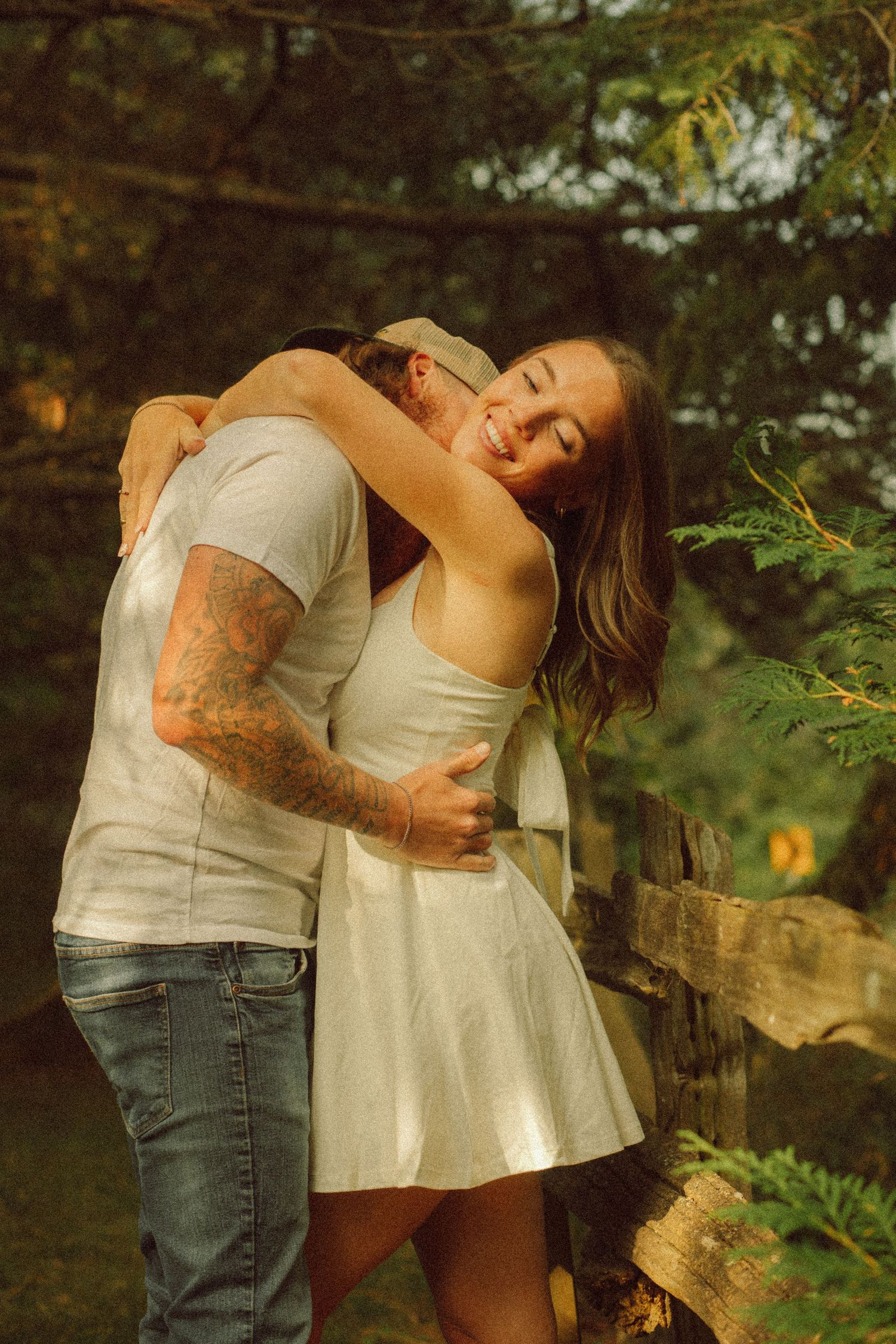 A couple embraces warmly outdoors, woman in white dress smiling joyfully, man with tattoos in jeans, surrounded by lush greenery.
