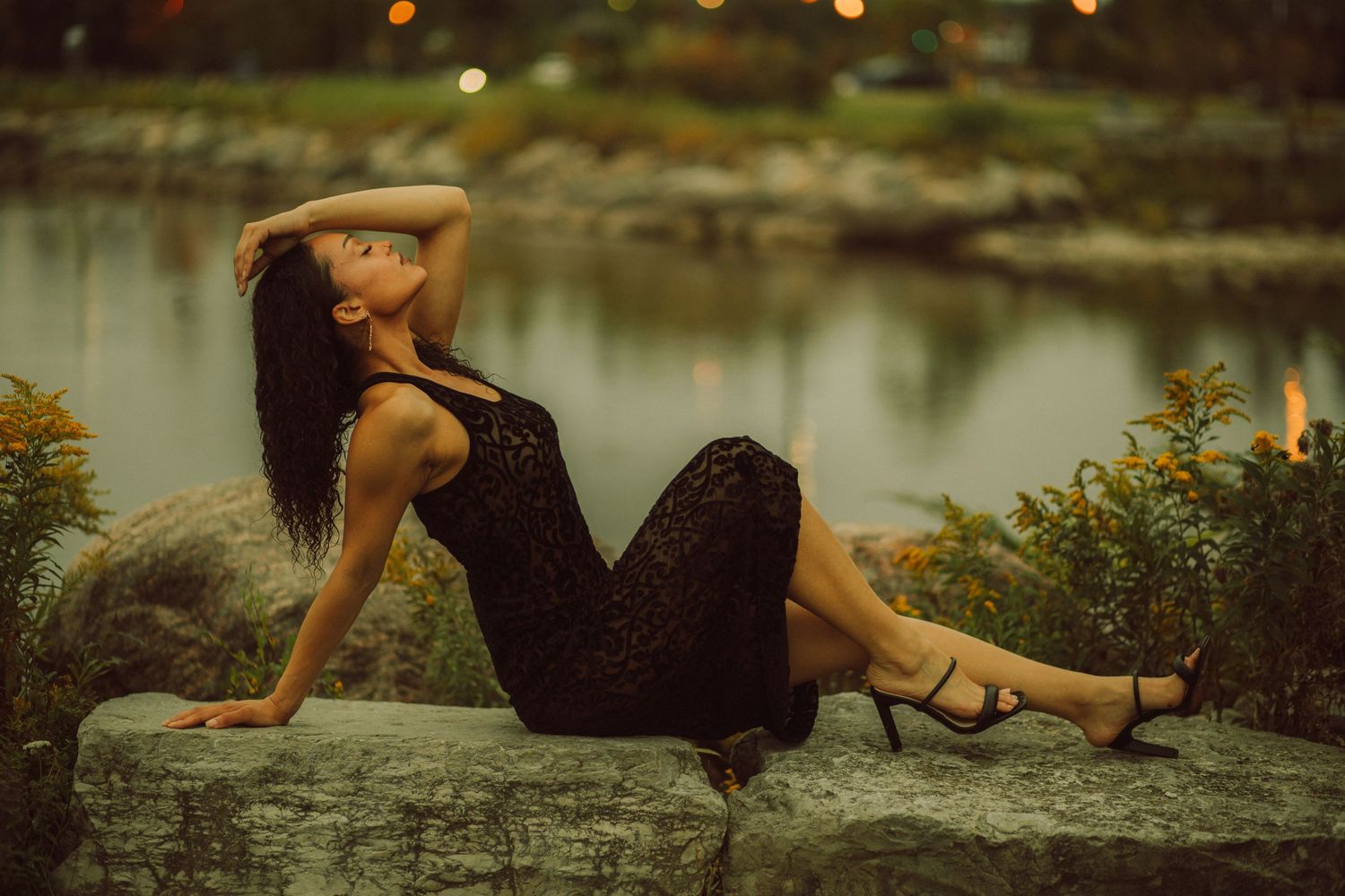 A woman in a black lace dress poses gracefully on rocks near a serene lake at dusk.