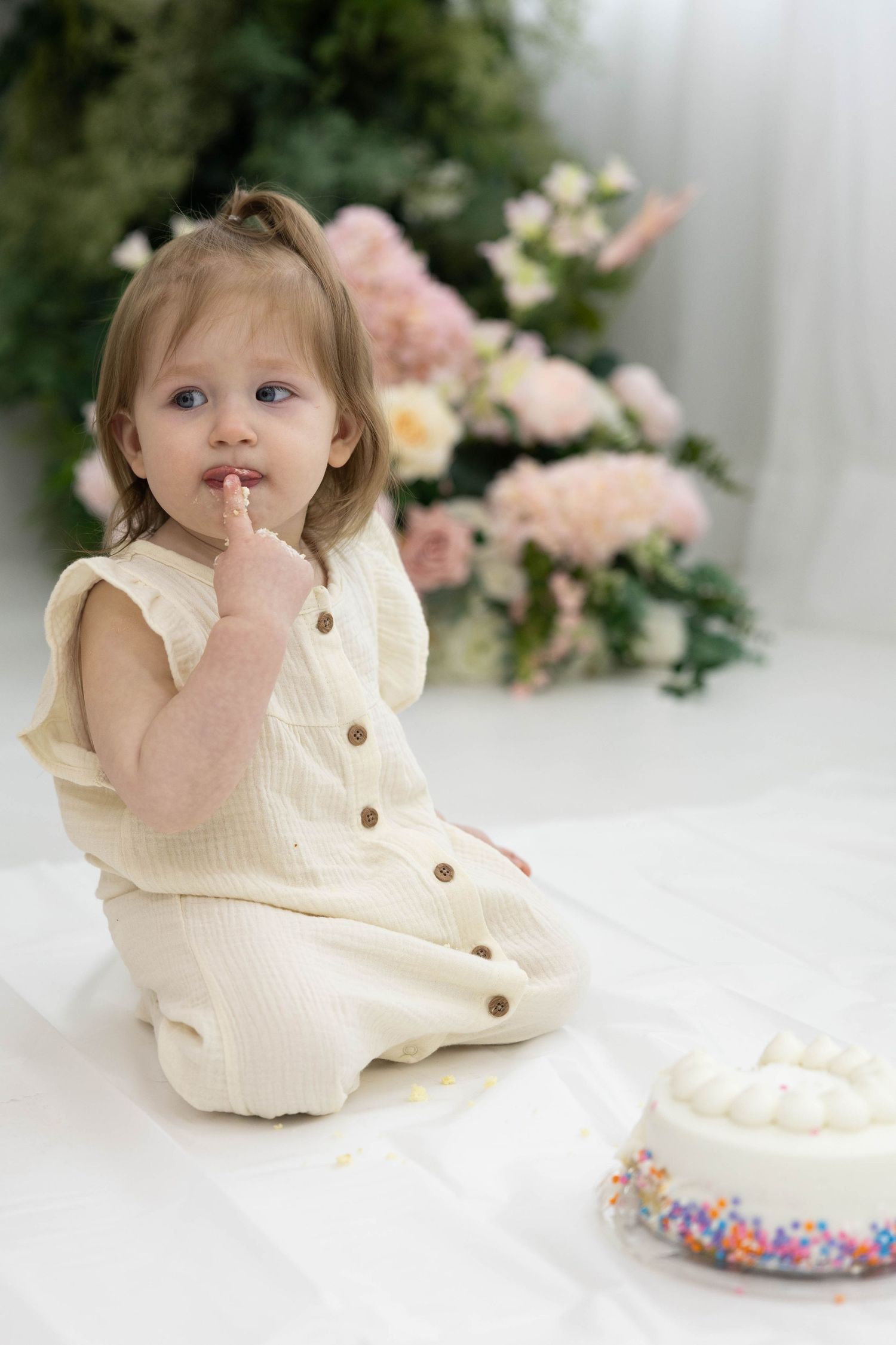 Small child in cream romper sitting near a white cake with sprinkles and floral backdrop.