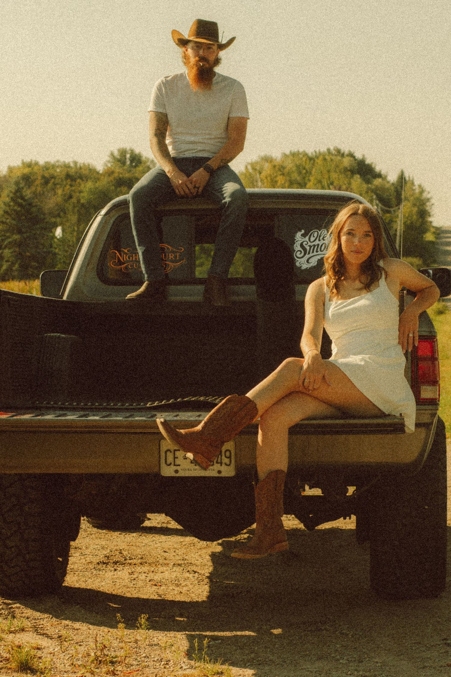 Man in cowboy hat and woman in white dress with cowboy boots posing on a pickup truck tailgate in a rural setting.