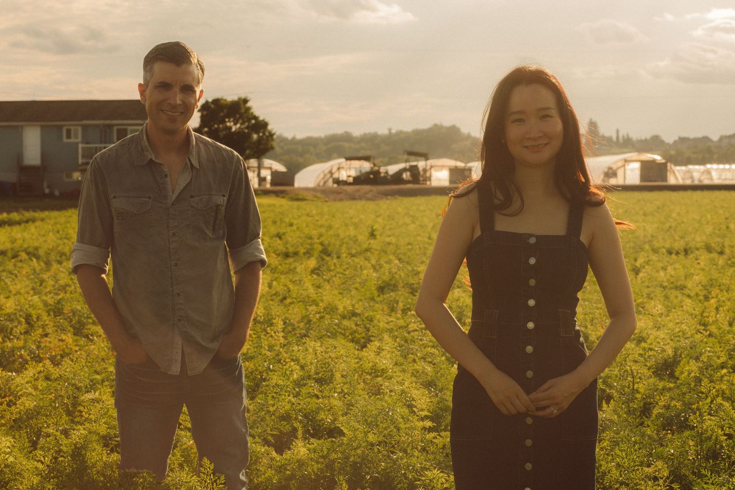 A man and woman stand in a lush green field at golden hour near farm buildings.