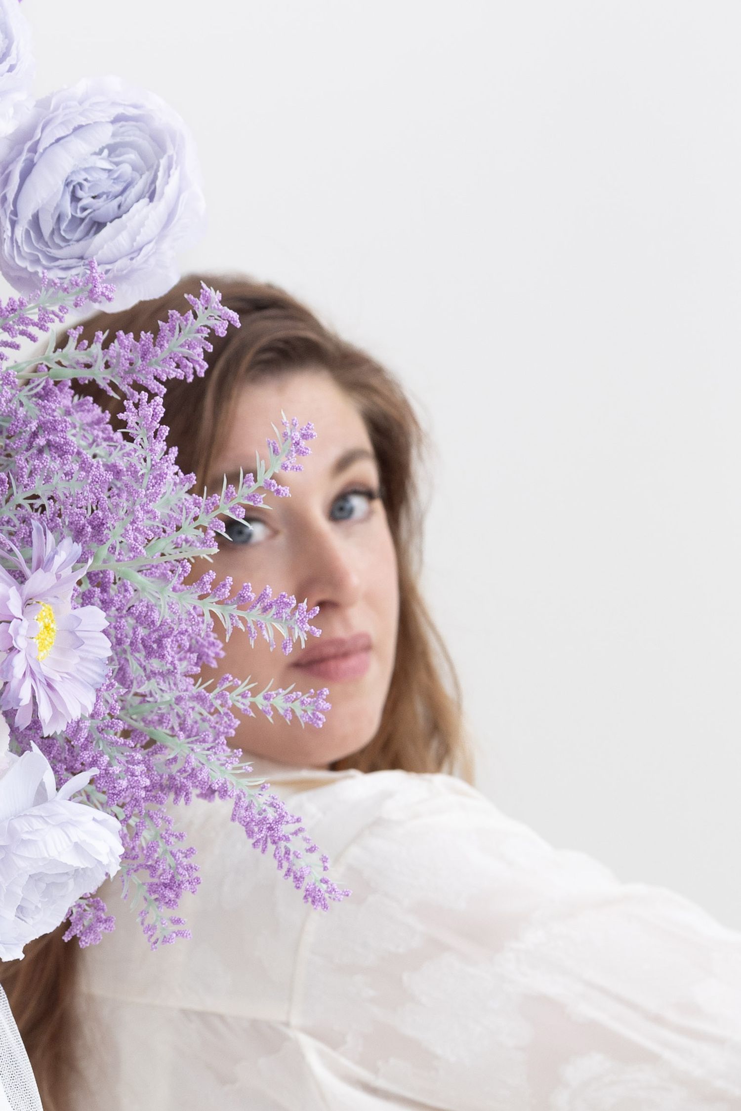 Woman partially hidden behind purple lavender and white roses bouquet wearing white dress.