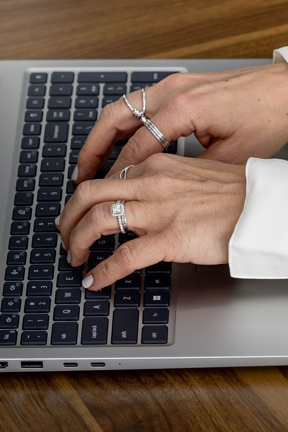 Woman's hands with diamond rings typing on a silver laptop keyboard on a wooden desk.