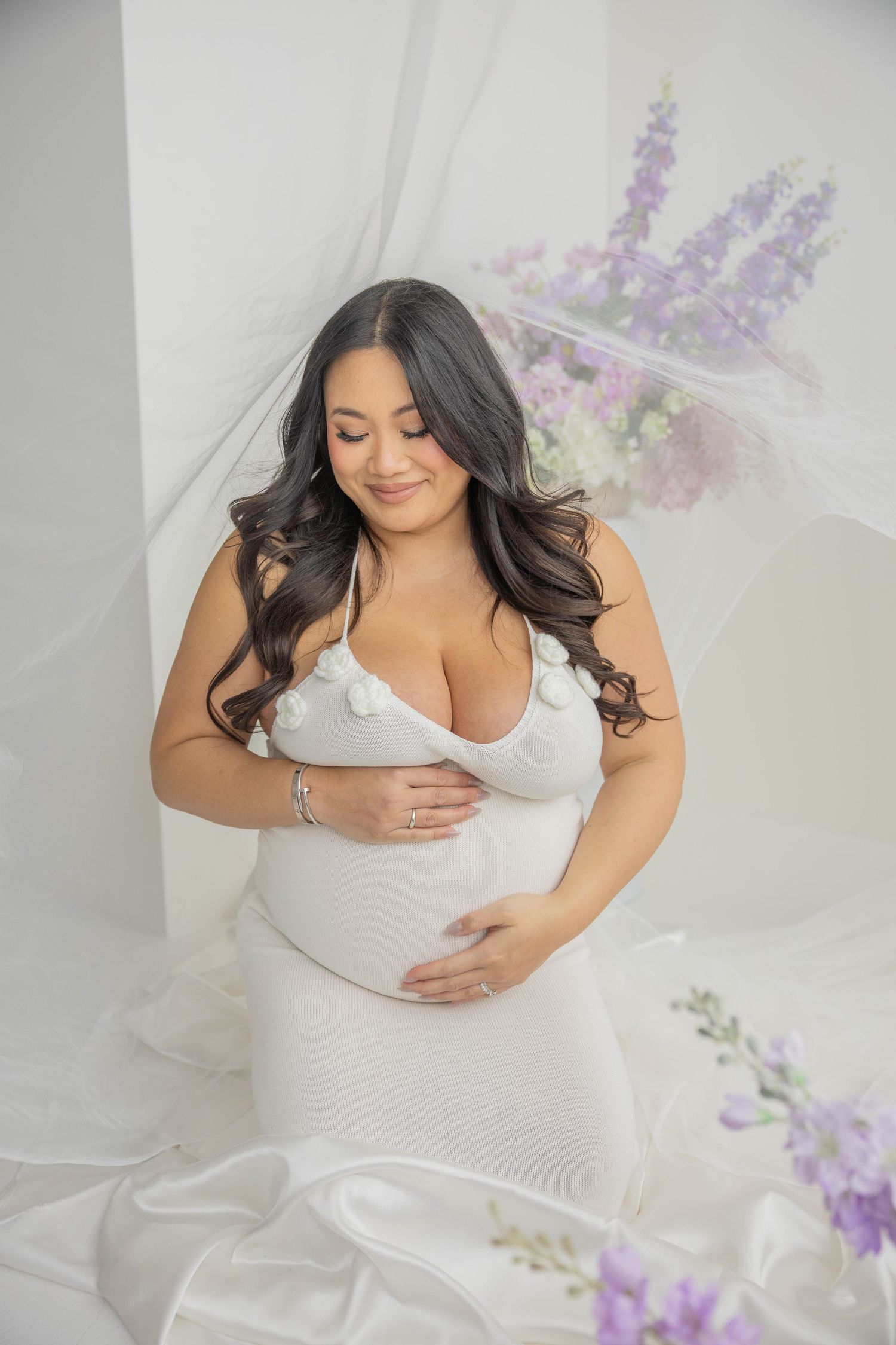 Intimate maternity session featuring white clothing and purple florals in a bright, airy studio space.