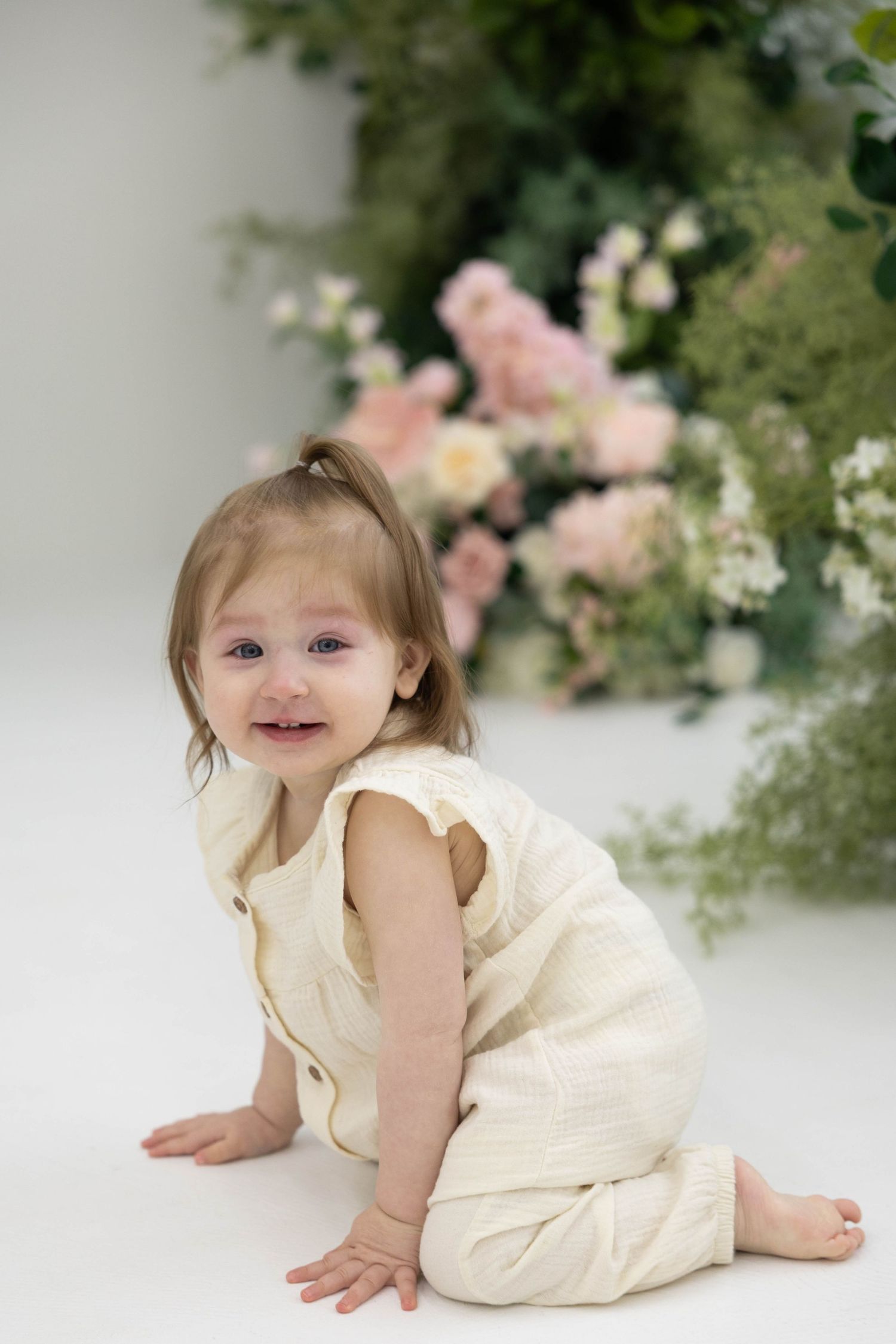 A small child in a cream-colored dress sits on a white floor with pink and white floral arrangements in the background.