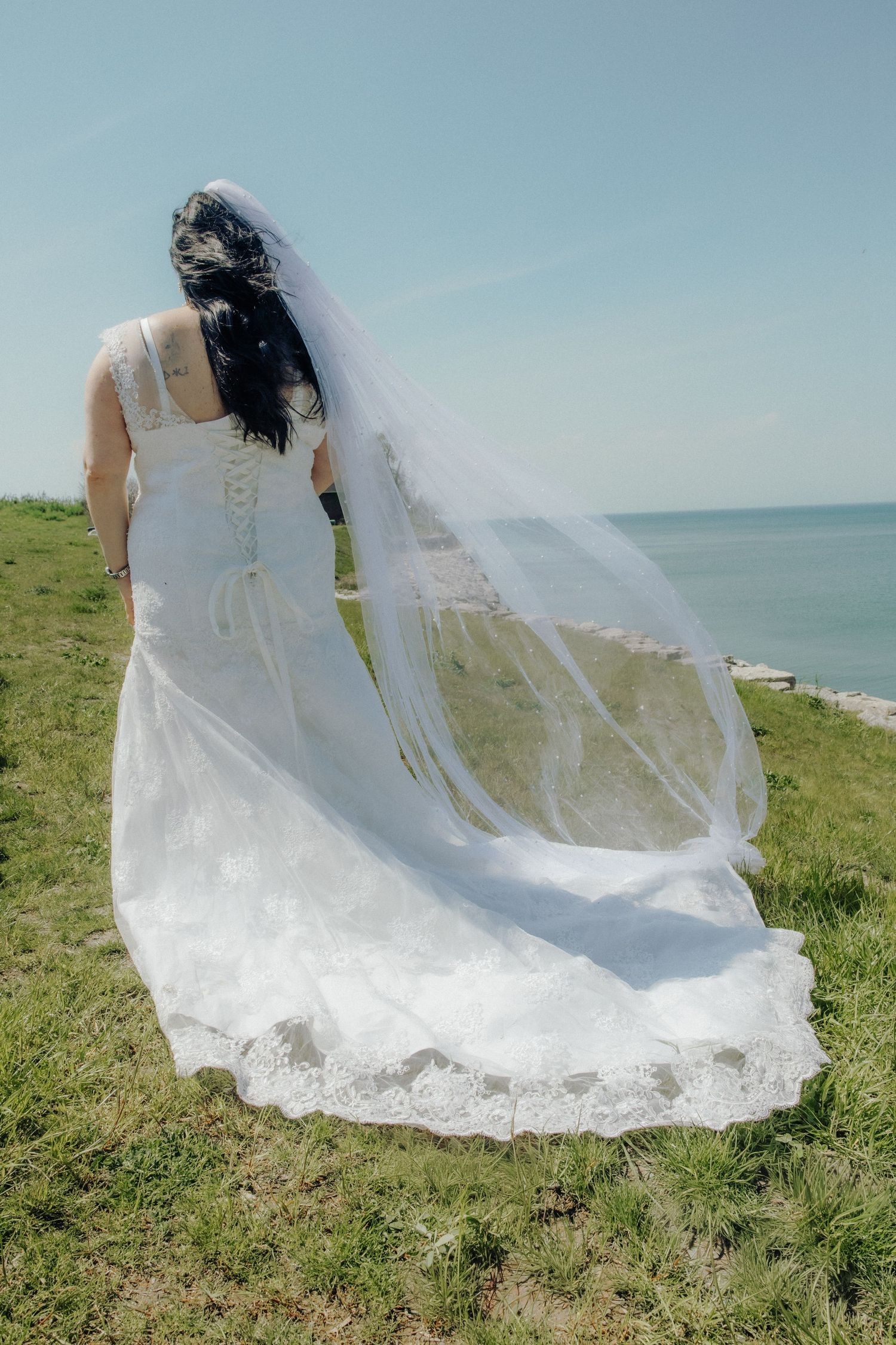 Bride in white lace wedding gown and flowing veil stands on green cliffside overlooking calm blue ocean on sunny day.