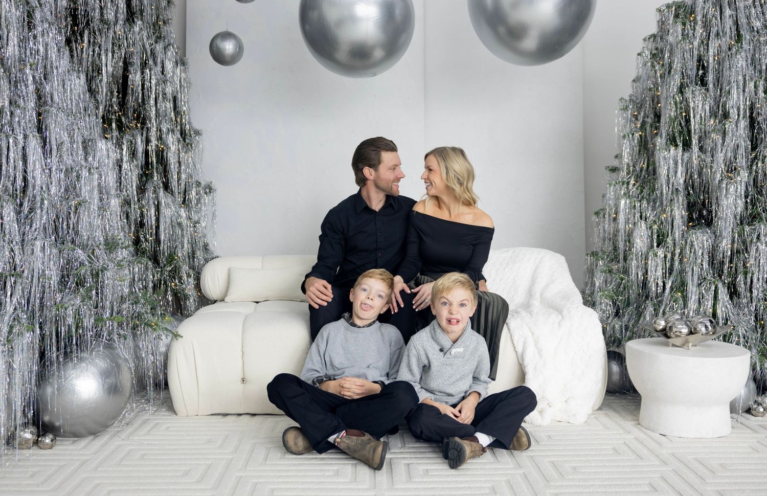Family of four posing for festive Christmas photo with silver decorations and white couch.