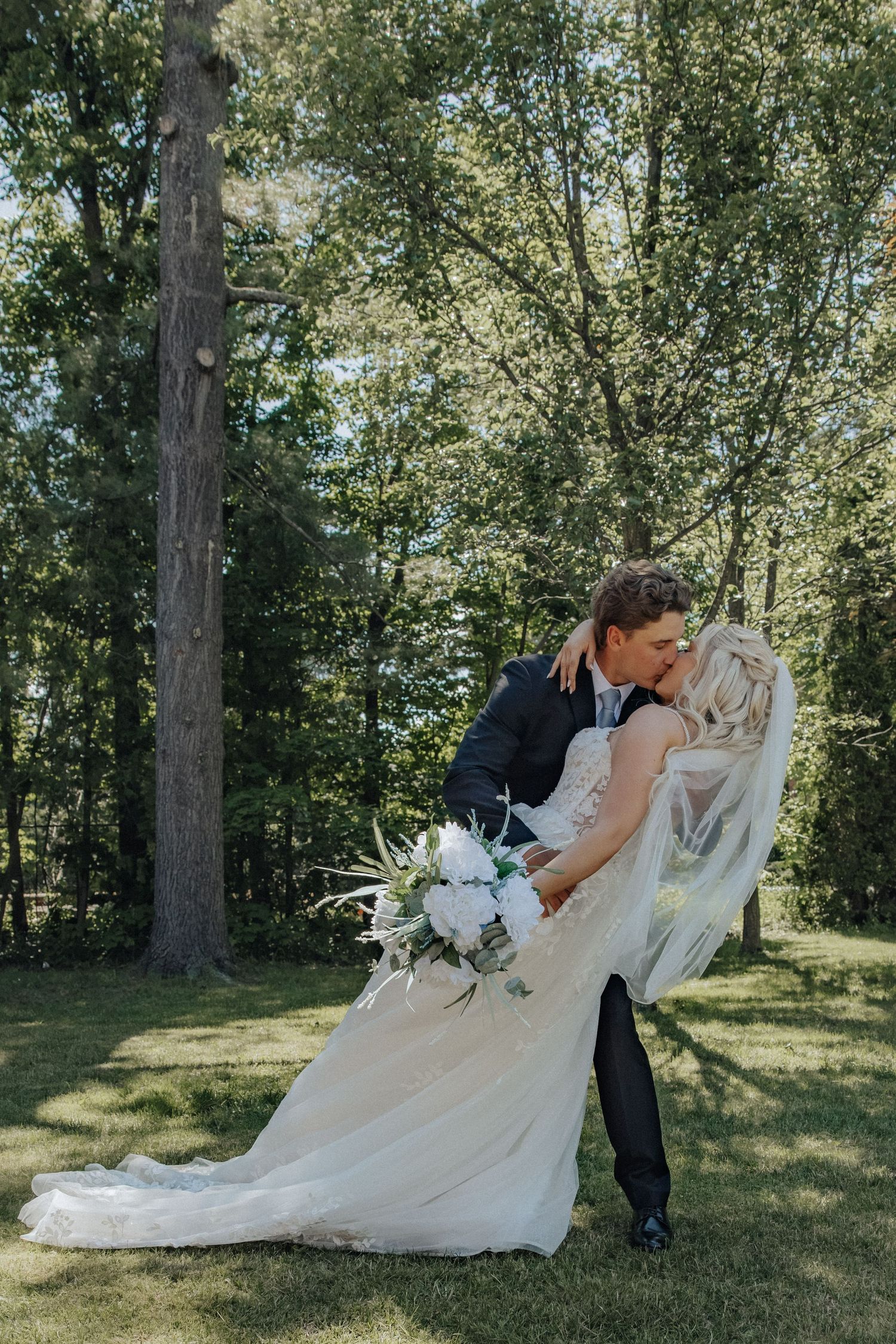 Groom dips bride for a kiss outdoors on green grass, surrounded by tall trees, bride holding white bouquet in wedding attire.