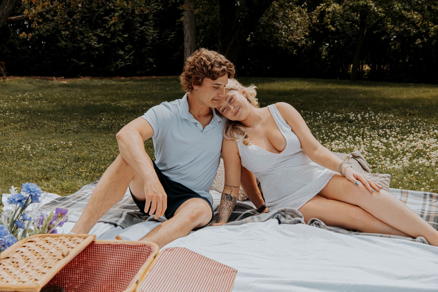 Romantic couple lying together on a blanket in a sunny park with white wildflowers.