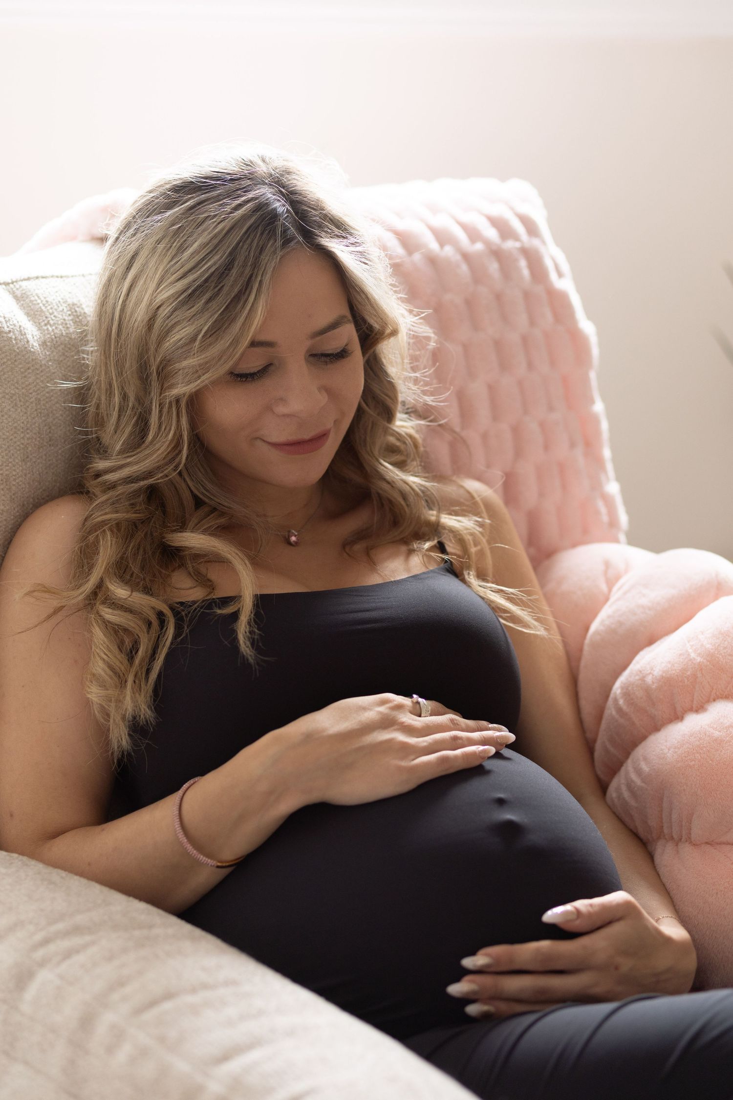 Pregnant woman in black dress sitting on couch, gently cradling her baby bump with a serene smile.