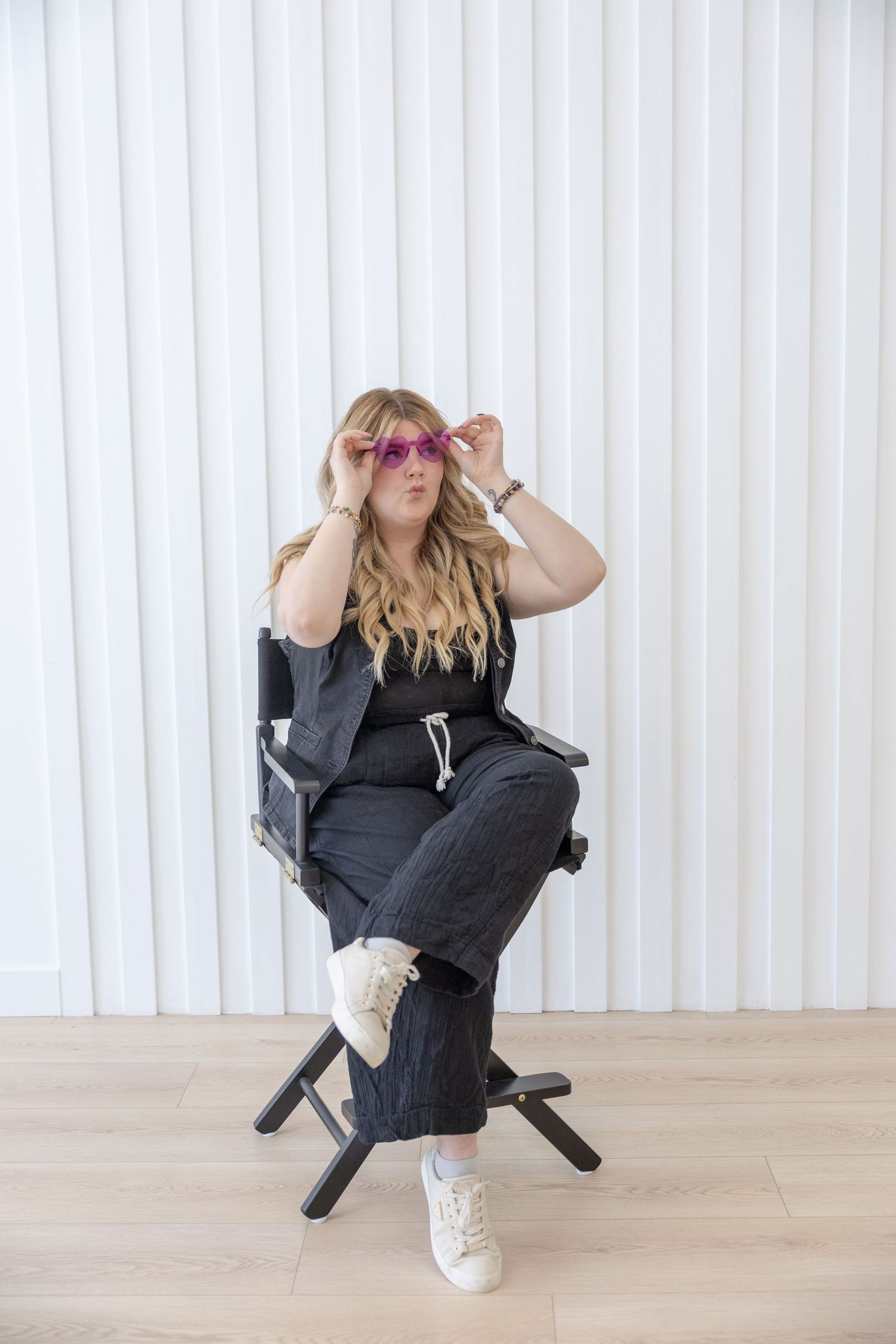 Woman in black outfit adjusting pink sunglasses while seated in a director's chair against a white wall.