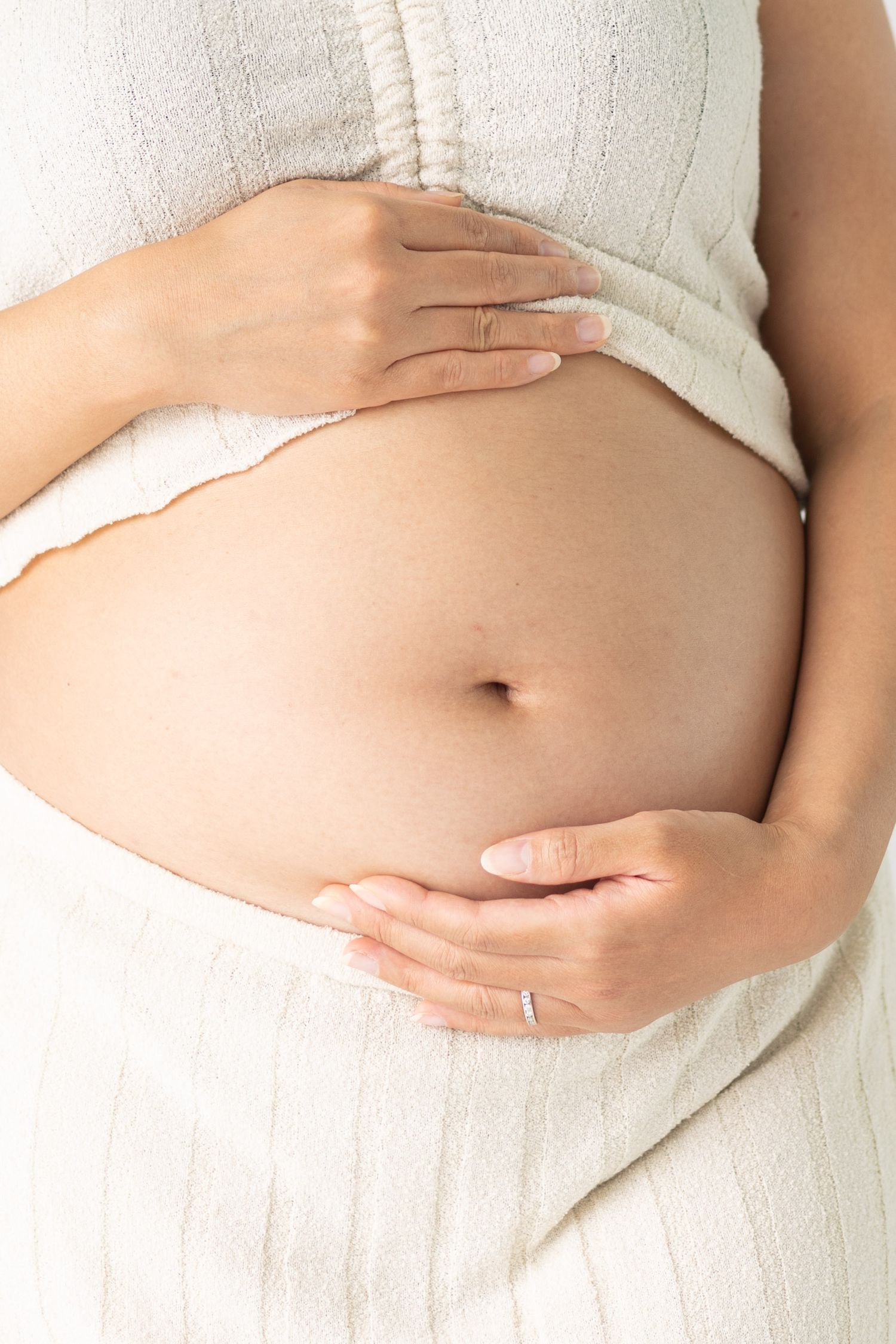 Pregnant woman in white knit dress cradling her bare baby bump with both hands, wearing a ring.