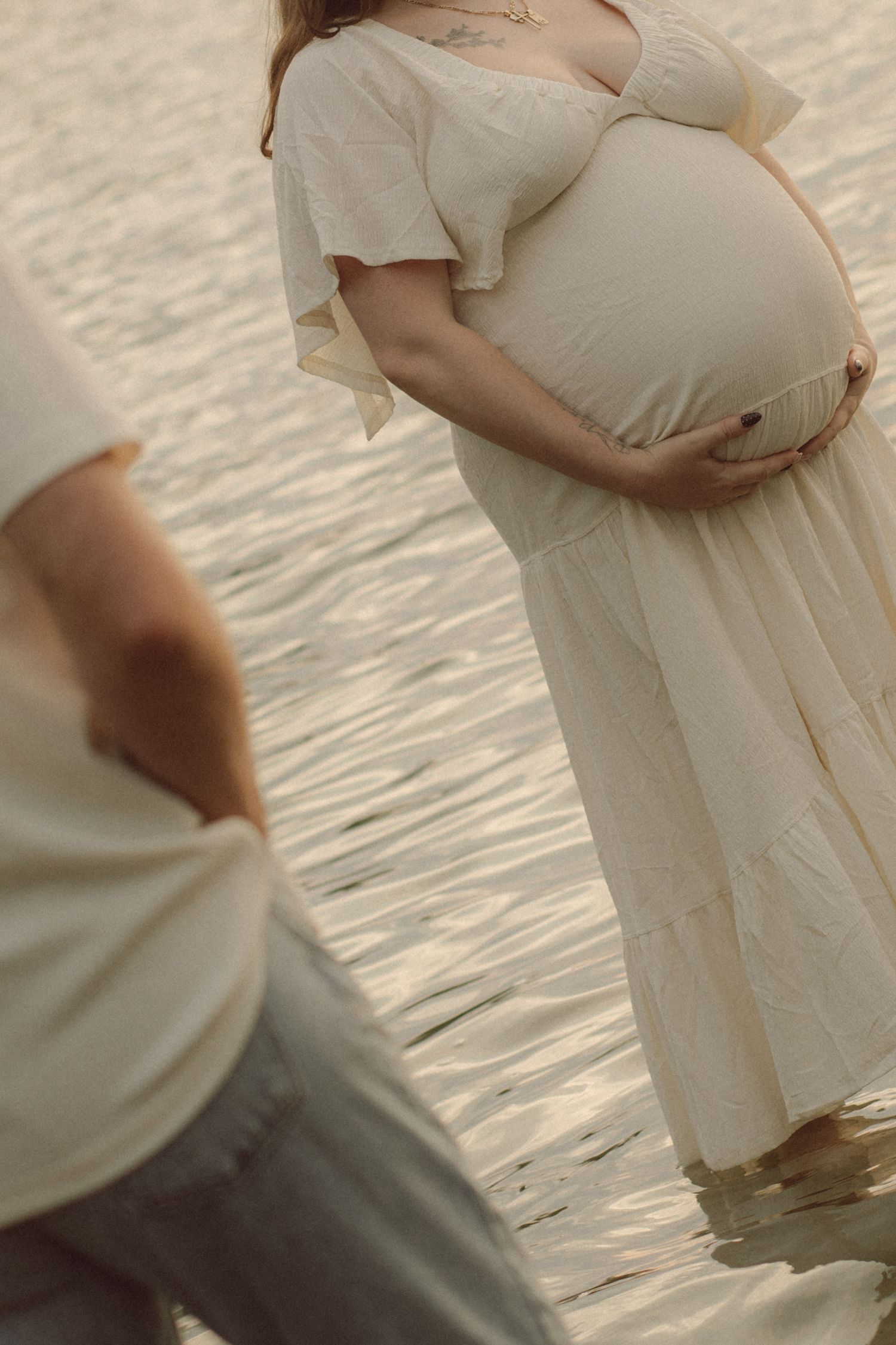 Pregnant woman in white dress cradling baby bump near water at golden hour during maternity photoshoot.
