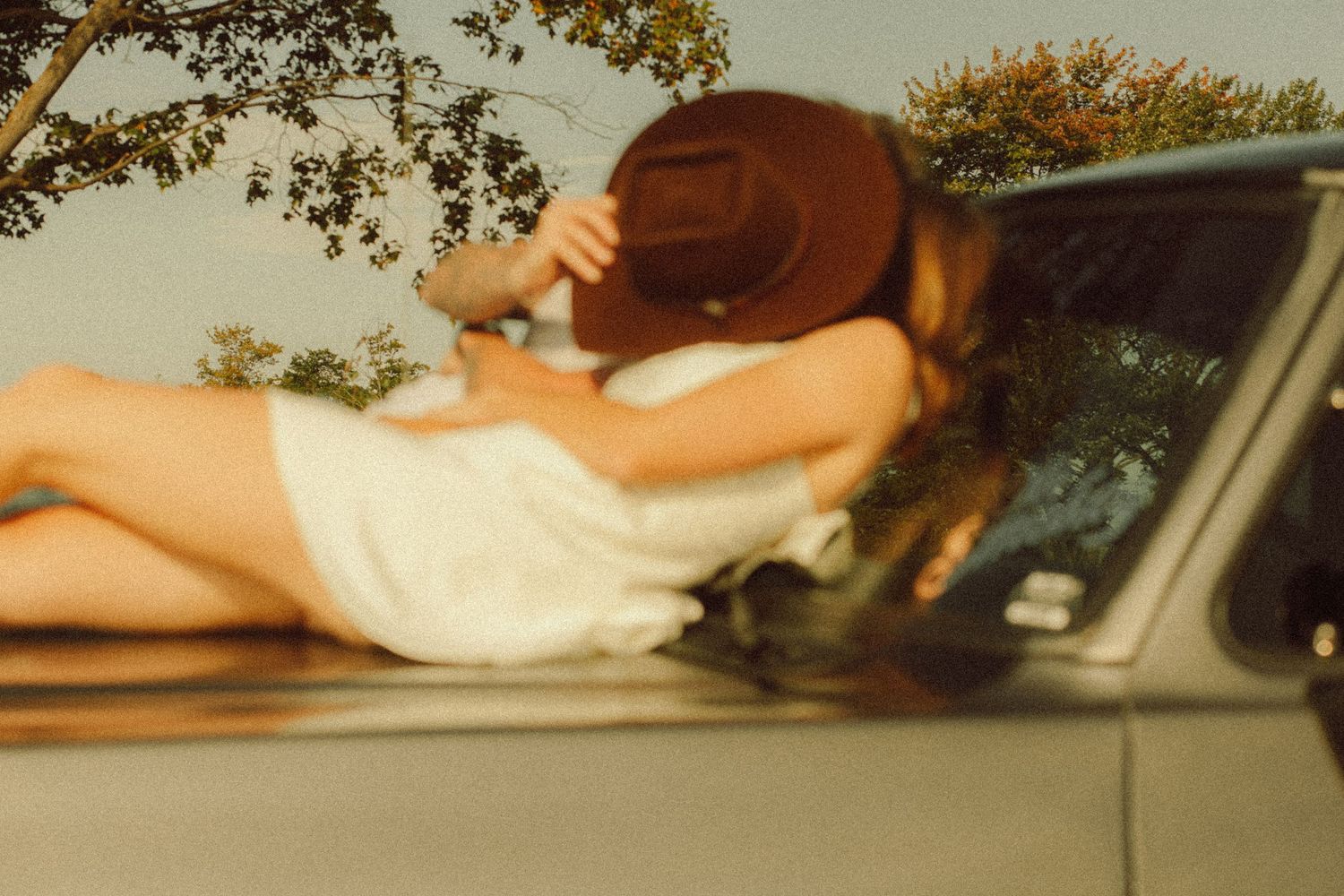 Woman in white dress and brown hat lying on vintage car hood outdoors.