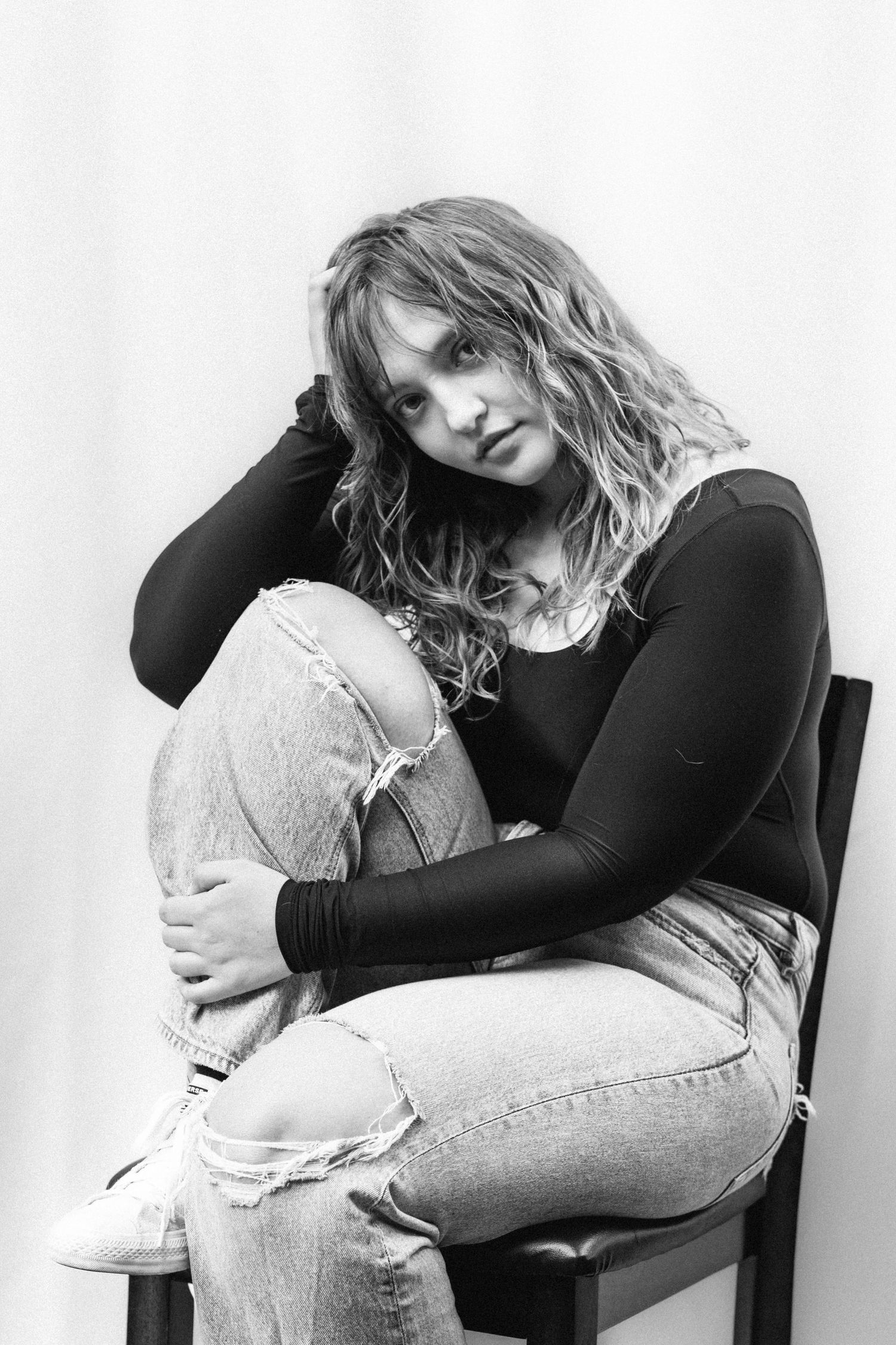 Black and white portrait of a young woman with wavy hair sitting on a chair, hugging her knees in ripped jeans.