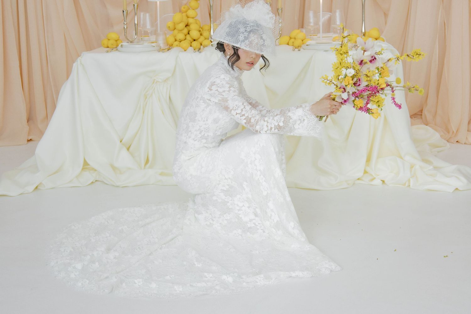 A bride in an elegant white lace wedding dress and headpiece arranges flowers at a decorated reception table.