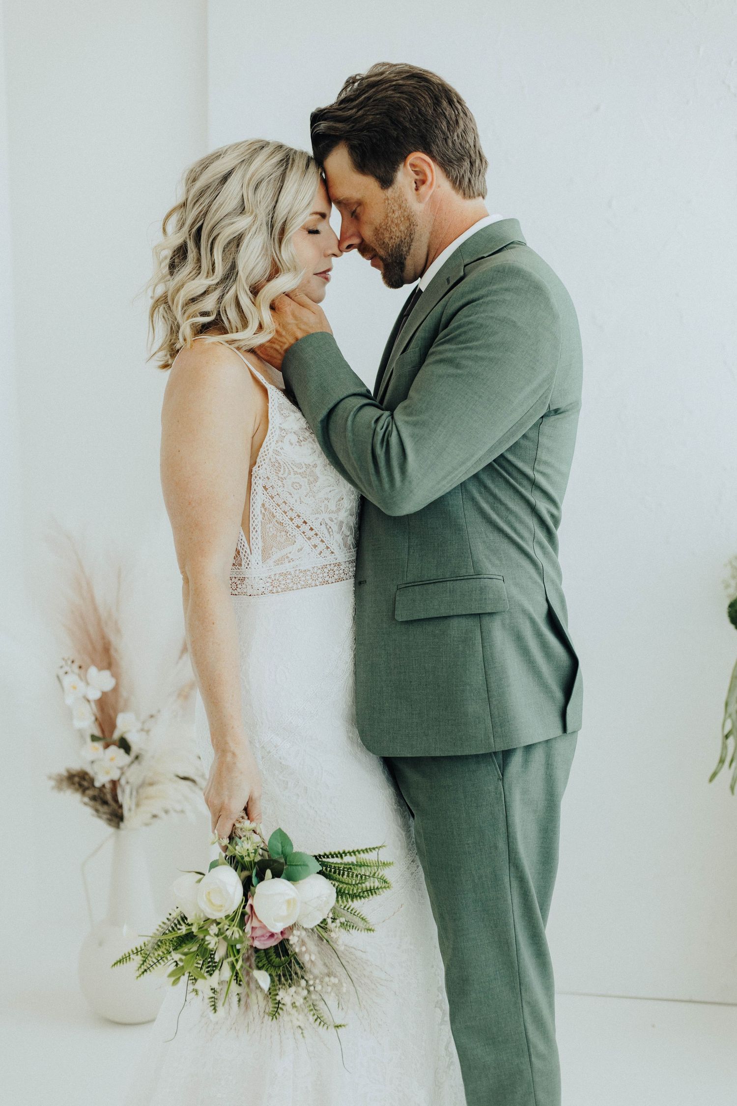 A romantic wedding portrait of a couple in an intimate pose with the bride in white lace and groom in sage green suit.