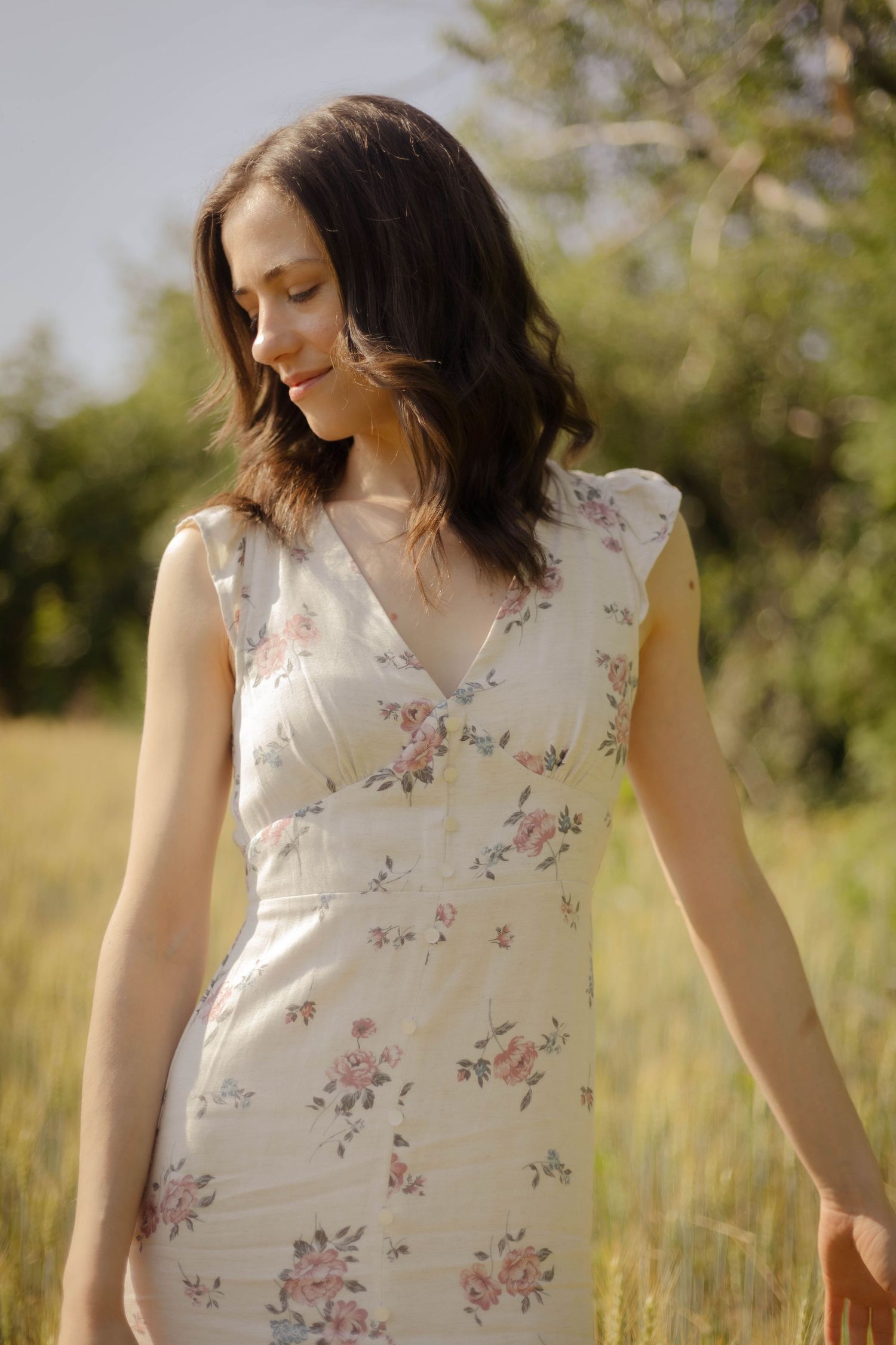 Young woman in white floral dress stands thoughtfully in a sunlit summer meadow with tall grass.