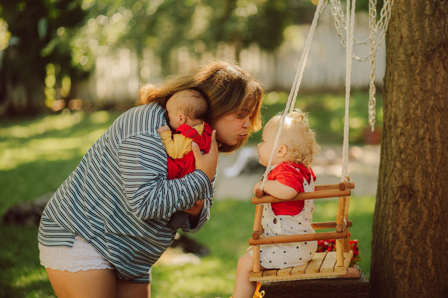 A mother holds her baby while leaning in to kiss a doll on a wooden swing outdoors.