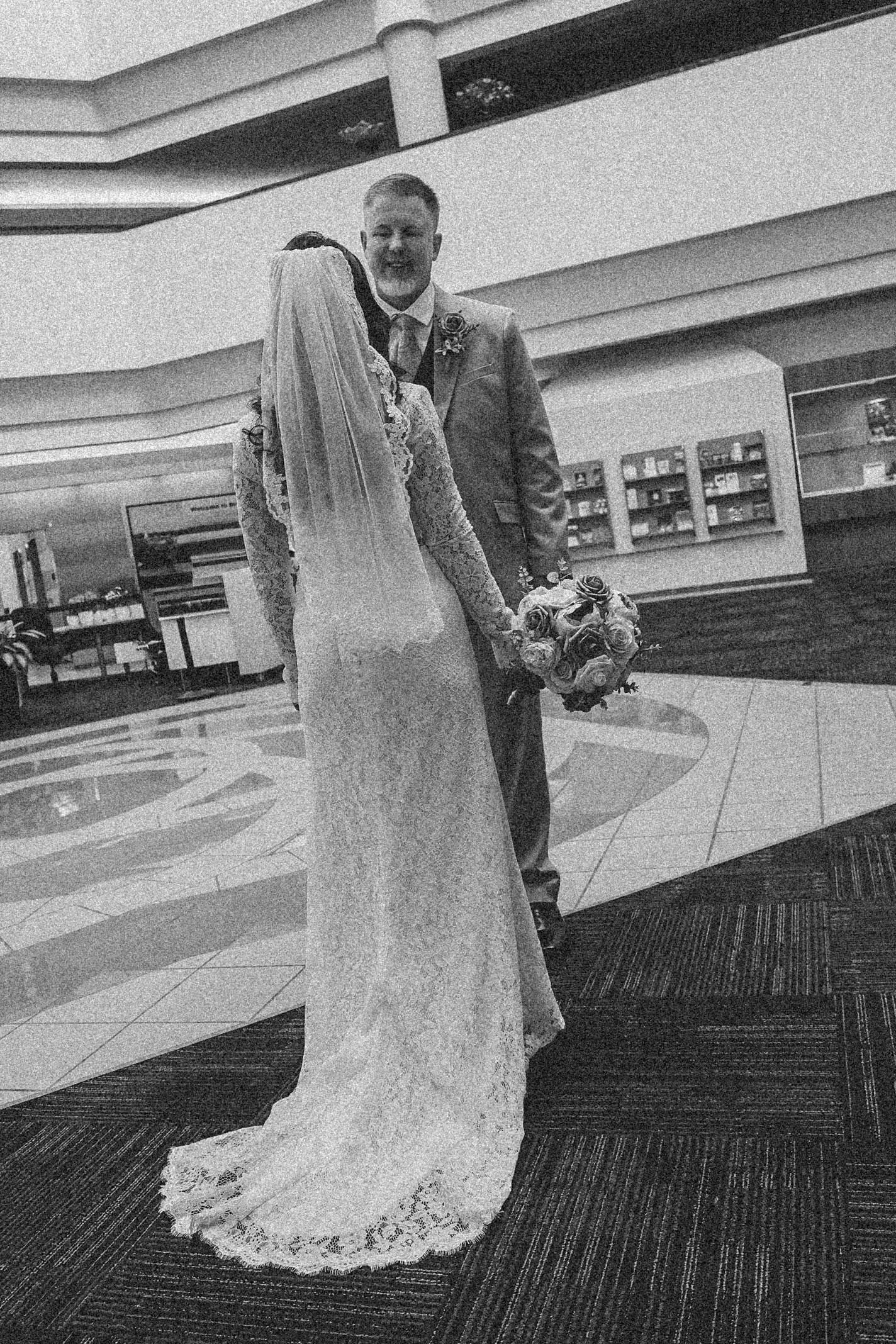A black and white wedding photo showing a couple embracing, with the bride wearing a long lace gown and holding a bouquet.