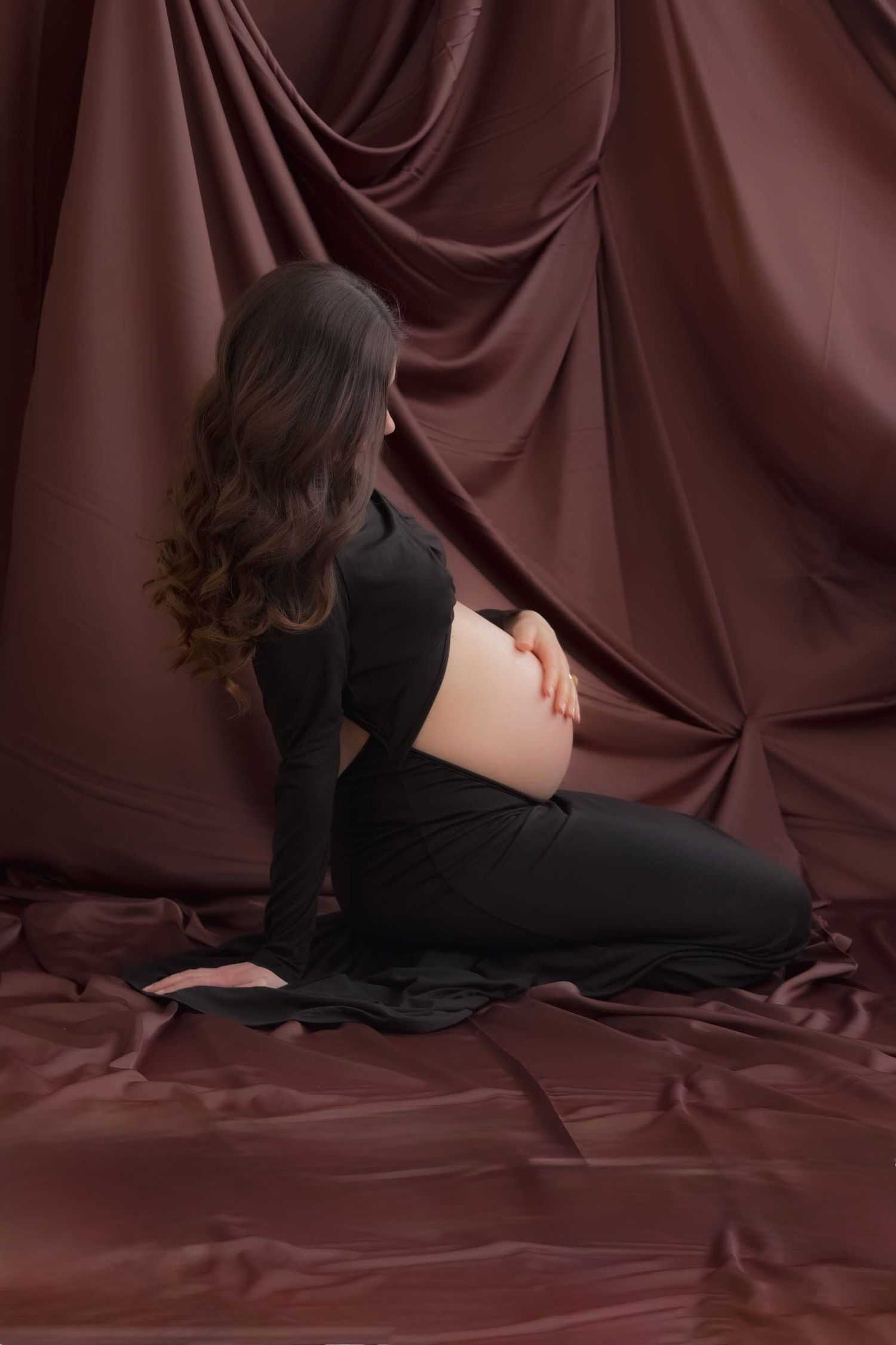 An artistic maternity portrait with burgundy backdrop and flowing black dress.
