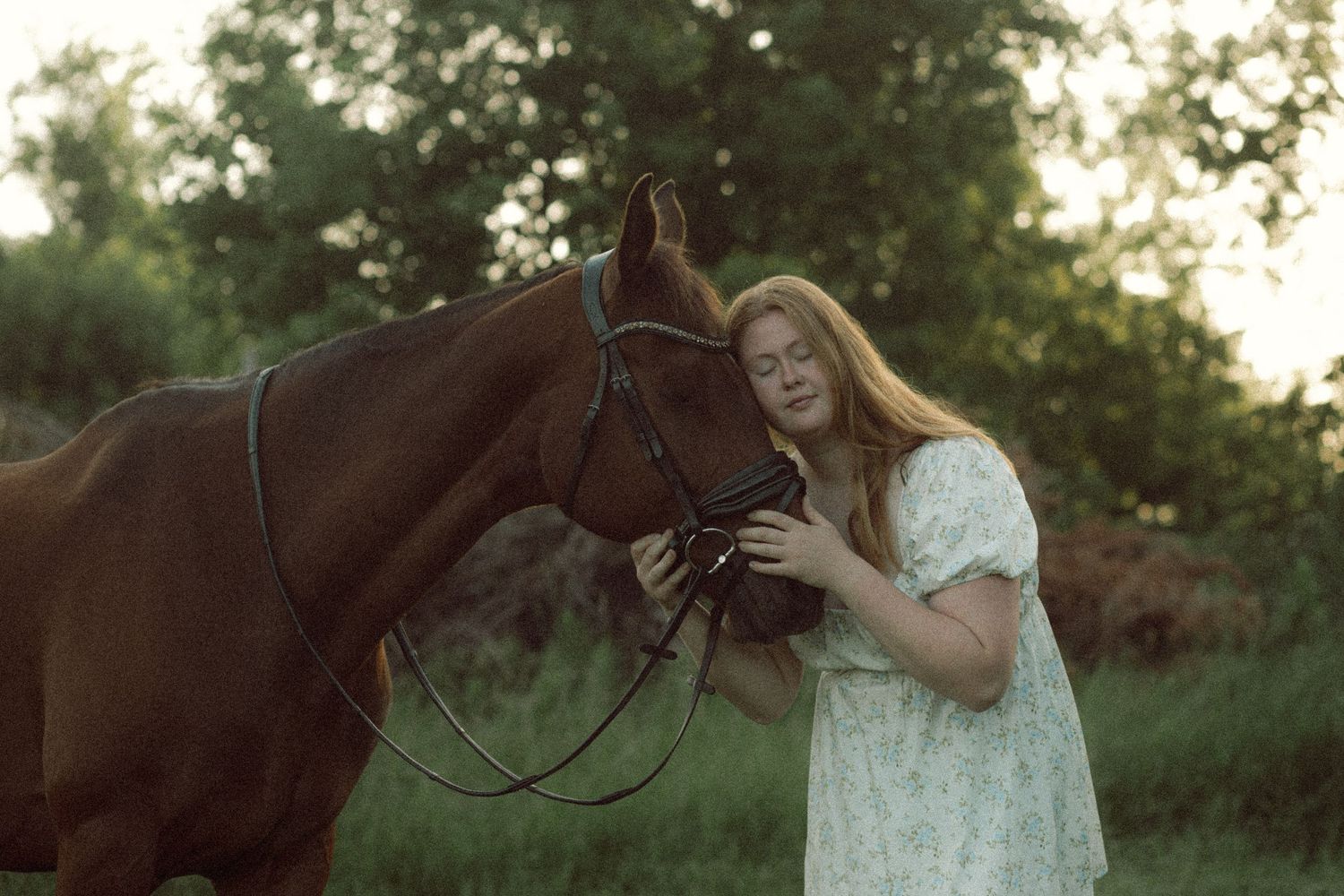 Young woman in white dress tenderly holding the bridle of a chestnut horse in a lush green outdoor setting.