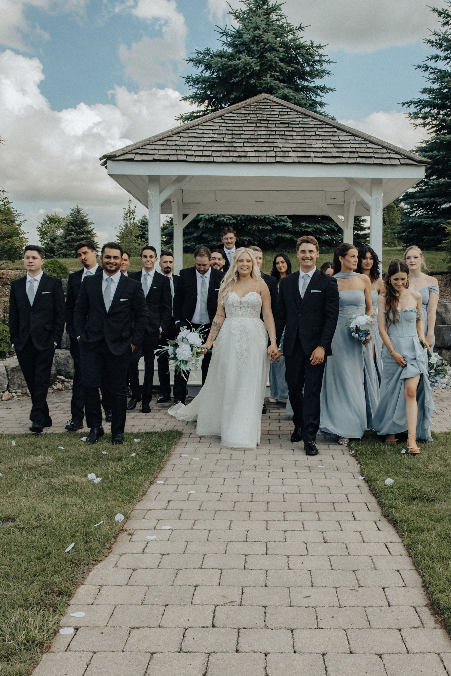 Wedding party poses on brick path before white gazebo, bride in white gown with groomsmen in suits and bridesmaids in blue dresses.
