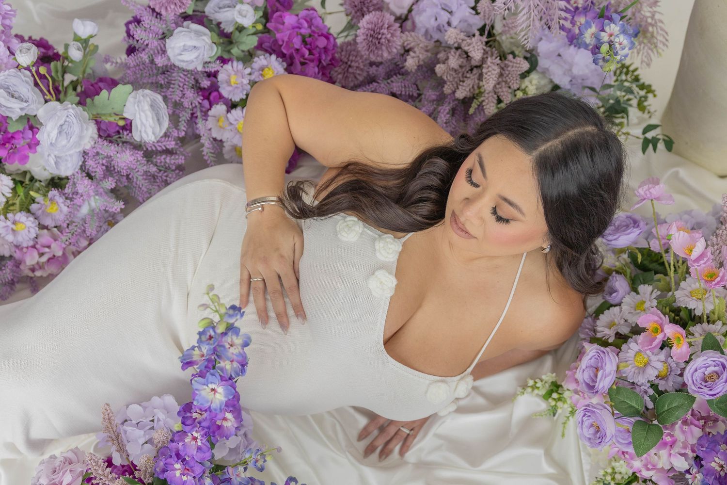A person in a white wedding dress lies surrounded by purple and pink flowers in a romantic styled photoshoot.