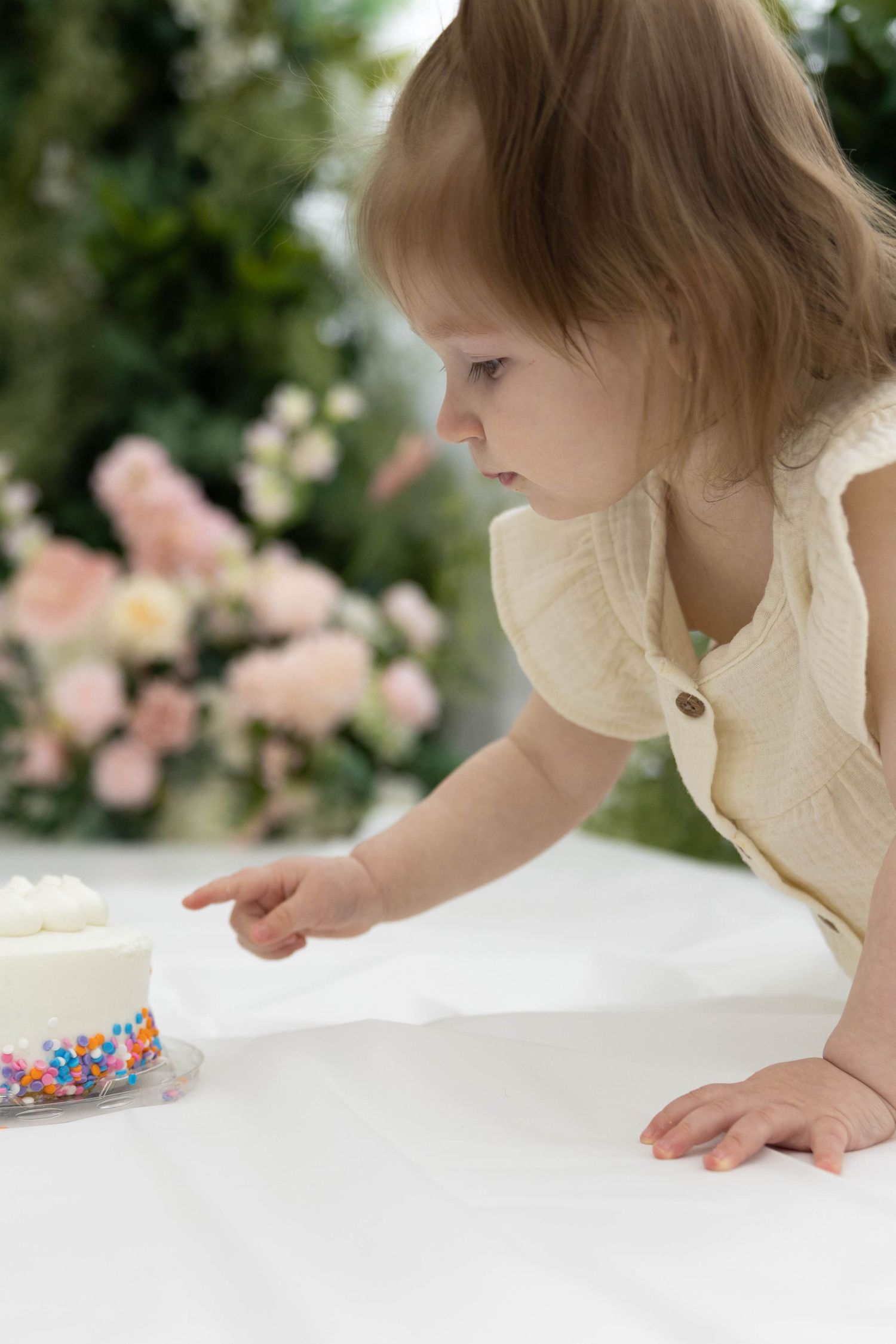 A small child in a ruffled cream outfit leans over to examine a decorated cake against a floral backdrop.