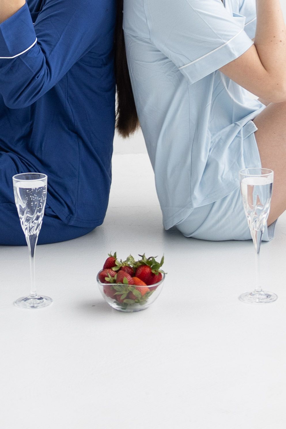 Two people in satin pajamas sitting back-to-back with champagne flutes and a bowl of fresh strawberries.