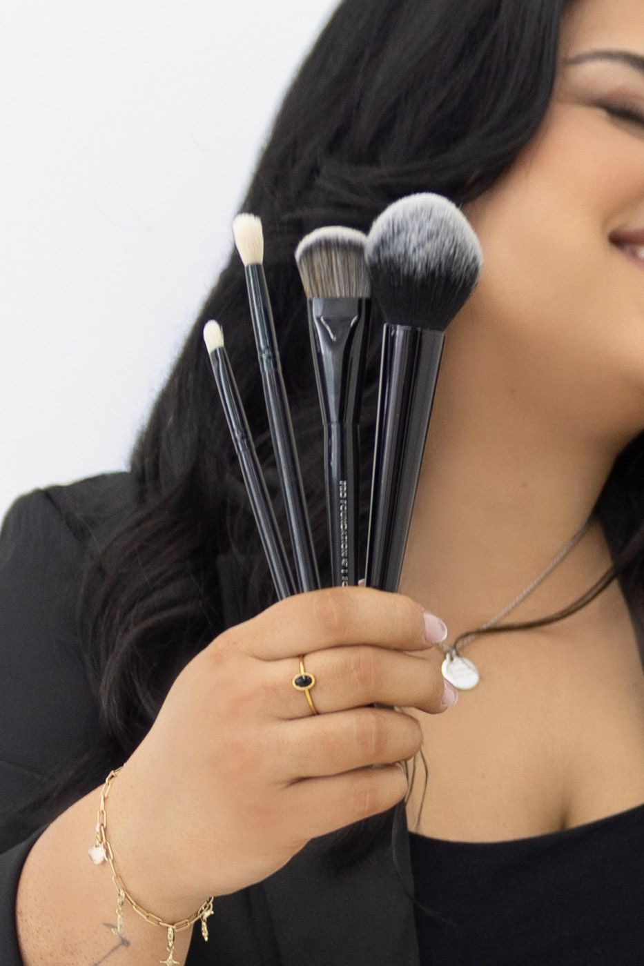 Woman holding a fan of five black-handled makeup brushes with soft bristles, wearing gold jewelry and a black outfit.