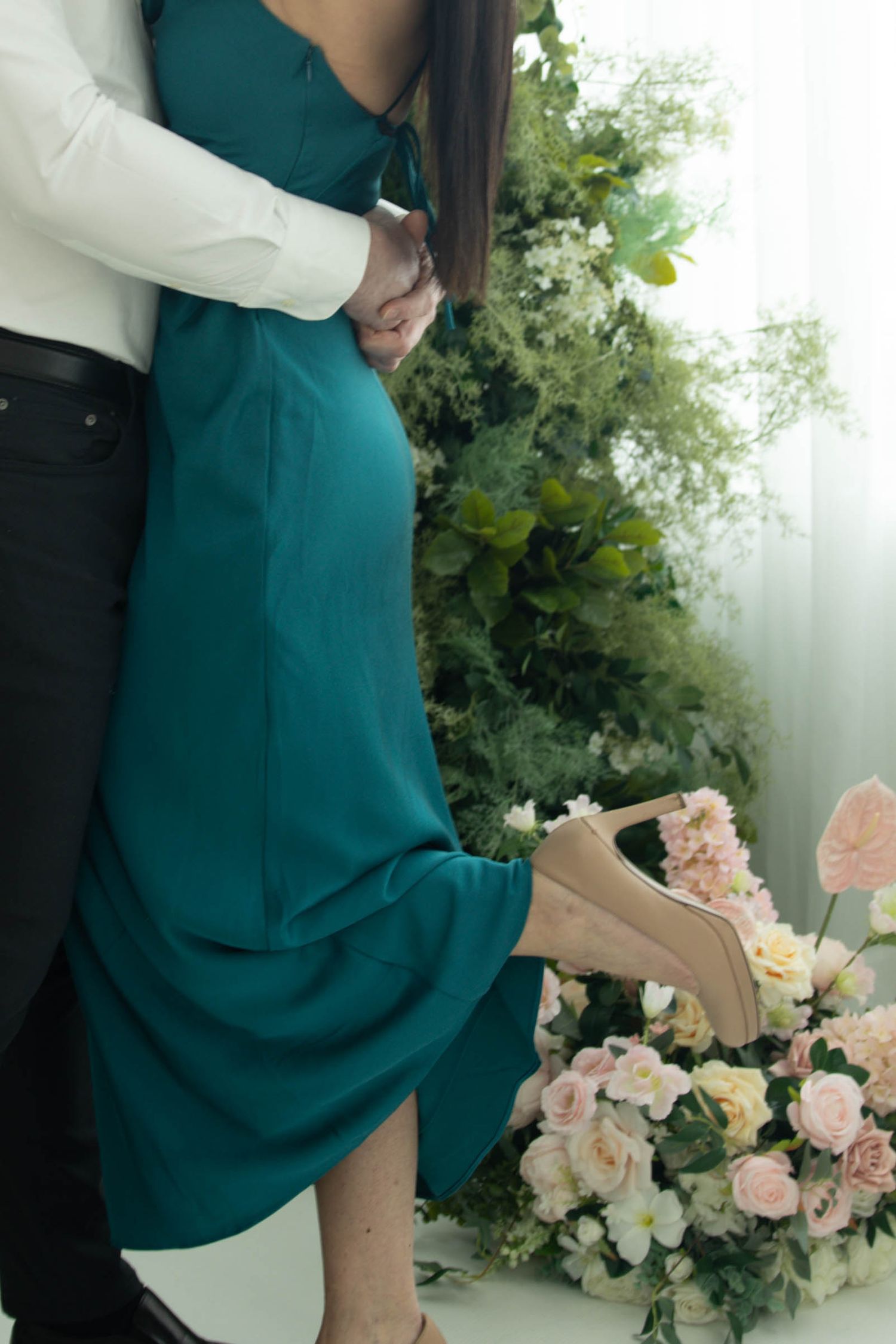 A romantic couple embraces in formal wear near floral arrangements featuring pink roses and white flowers in an outdoor setting.
