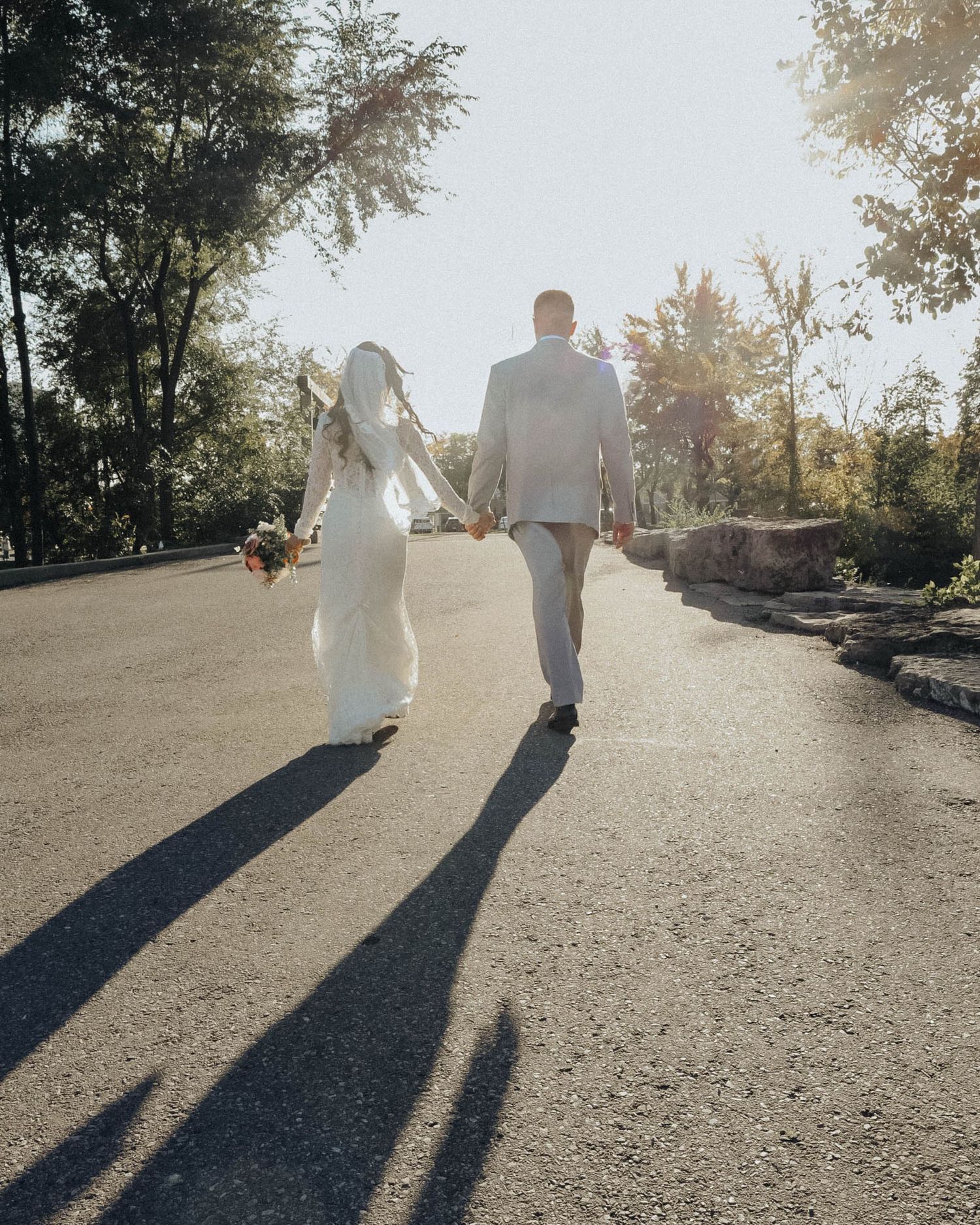 A couple in wedding attire walks down a sunlit path holding hands, casting long shadows behind them.