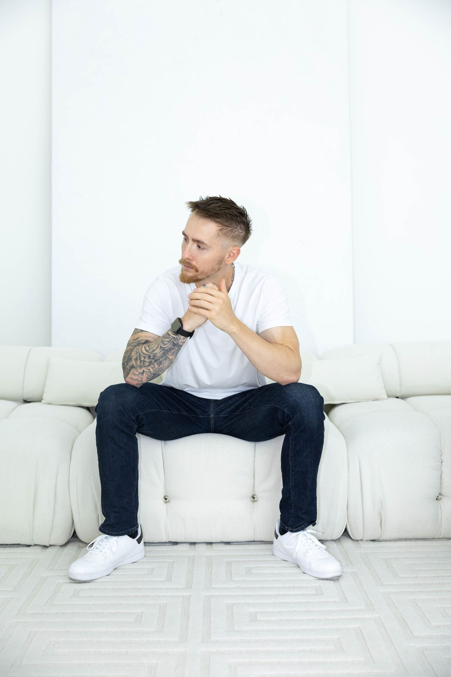 Person in white t-shirt and dark pants sits contemplatively on white couch in minimalist room.