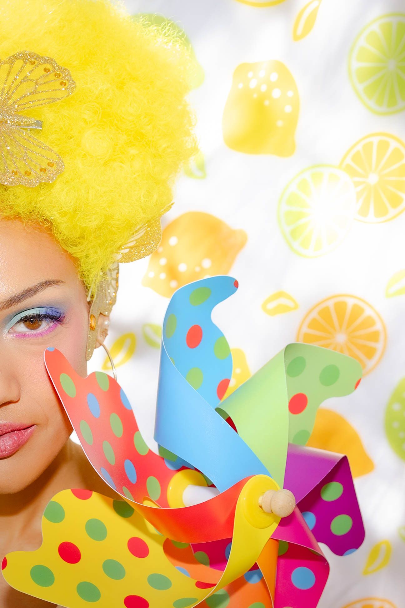 Vibrant artistic portrait with bright yellow hair and colorful pinwheel against citrus fruit background.