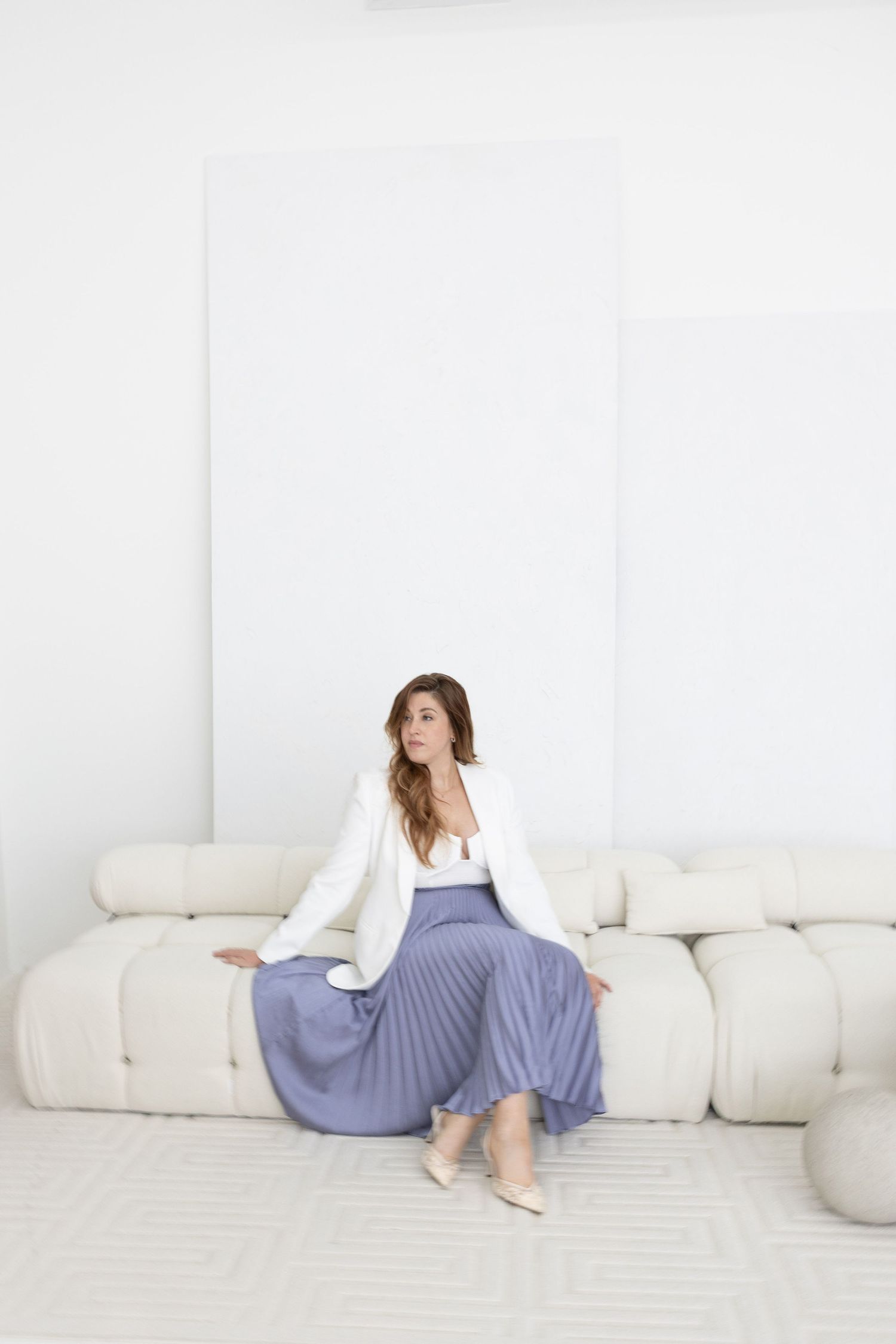 Stylish woman in white blazer and blue pleated skirt seated on modern white sofa in bright room.