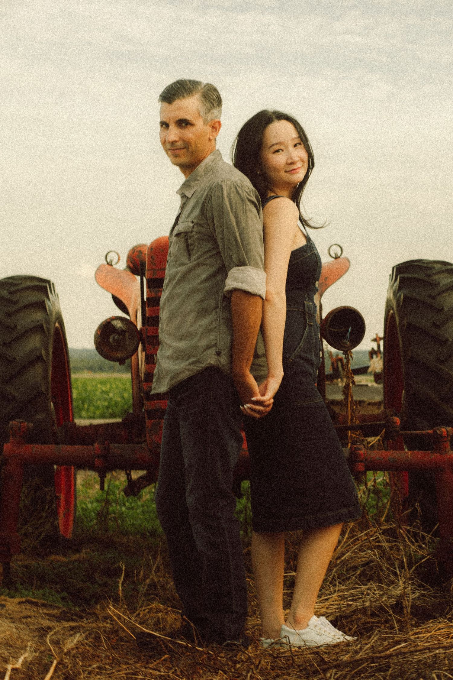 Couple standing back-to-back holding hands in front of a red tractor on a farm.