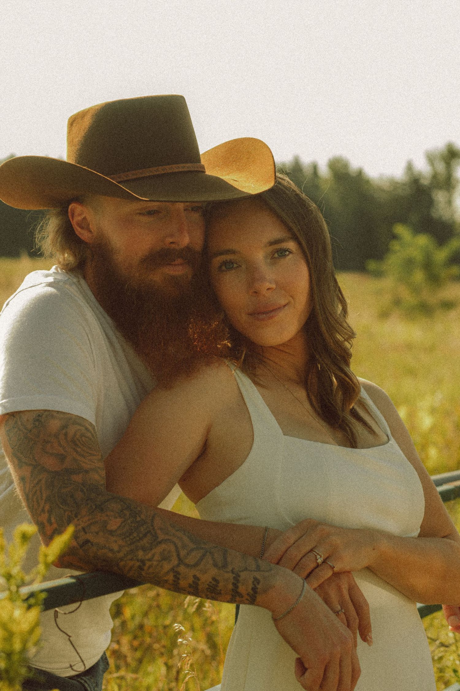 Tattooed bearded man in cowboy hat embracing brunette woman in white dress outdoors.