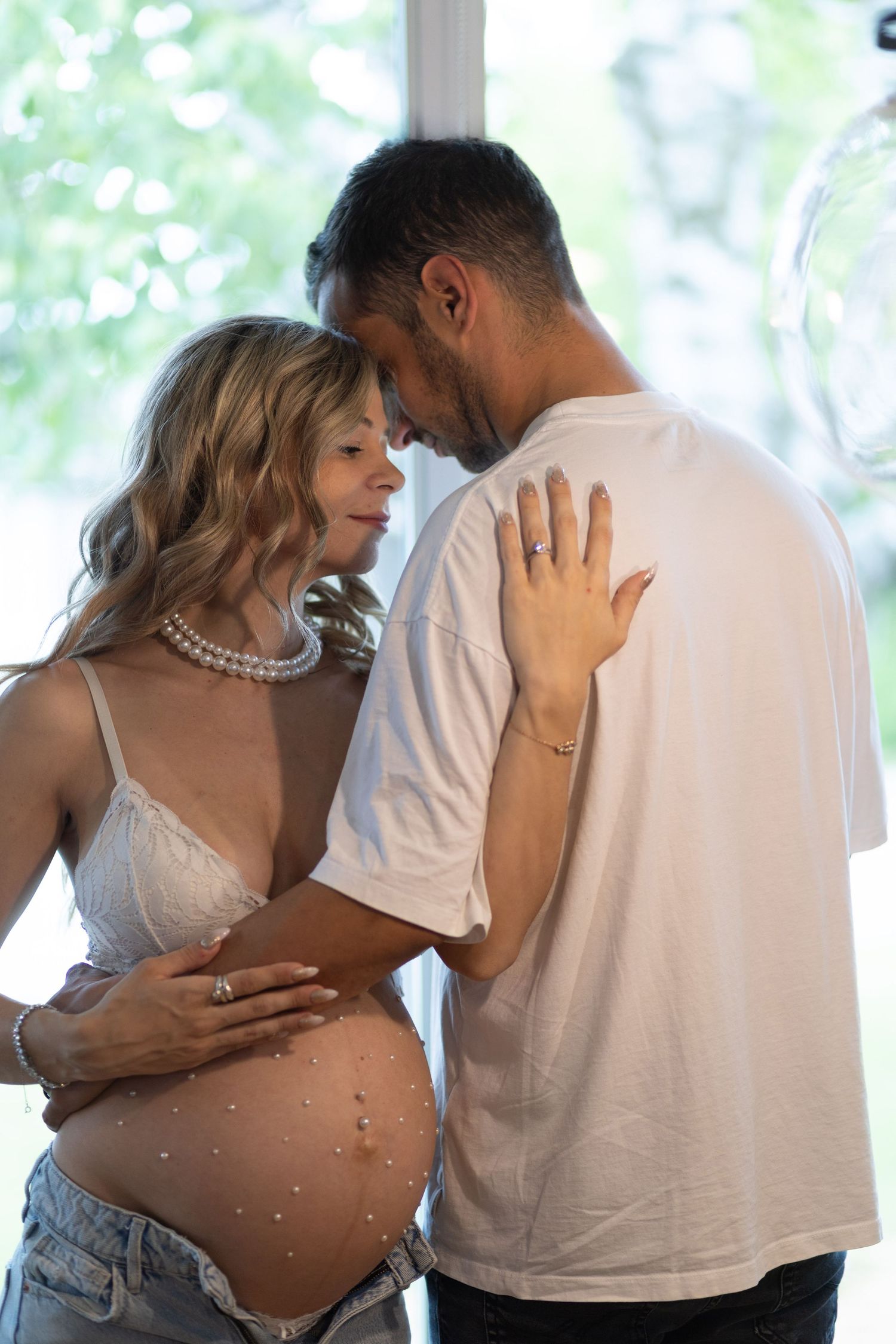 Expecting couple tenderly touching foreheads, pregnant woman adorned with pearls on belly.