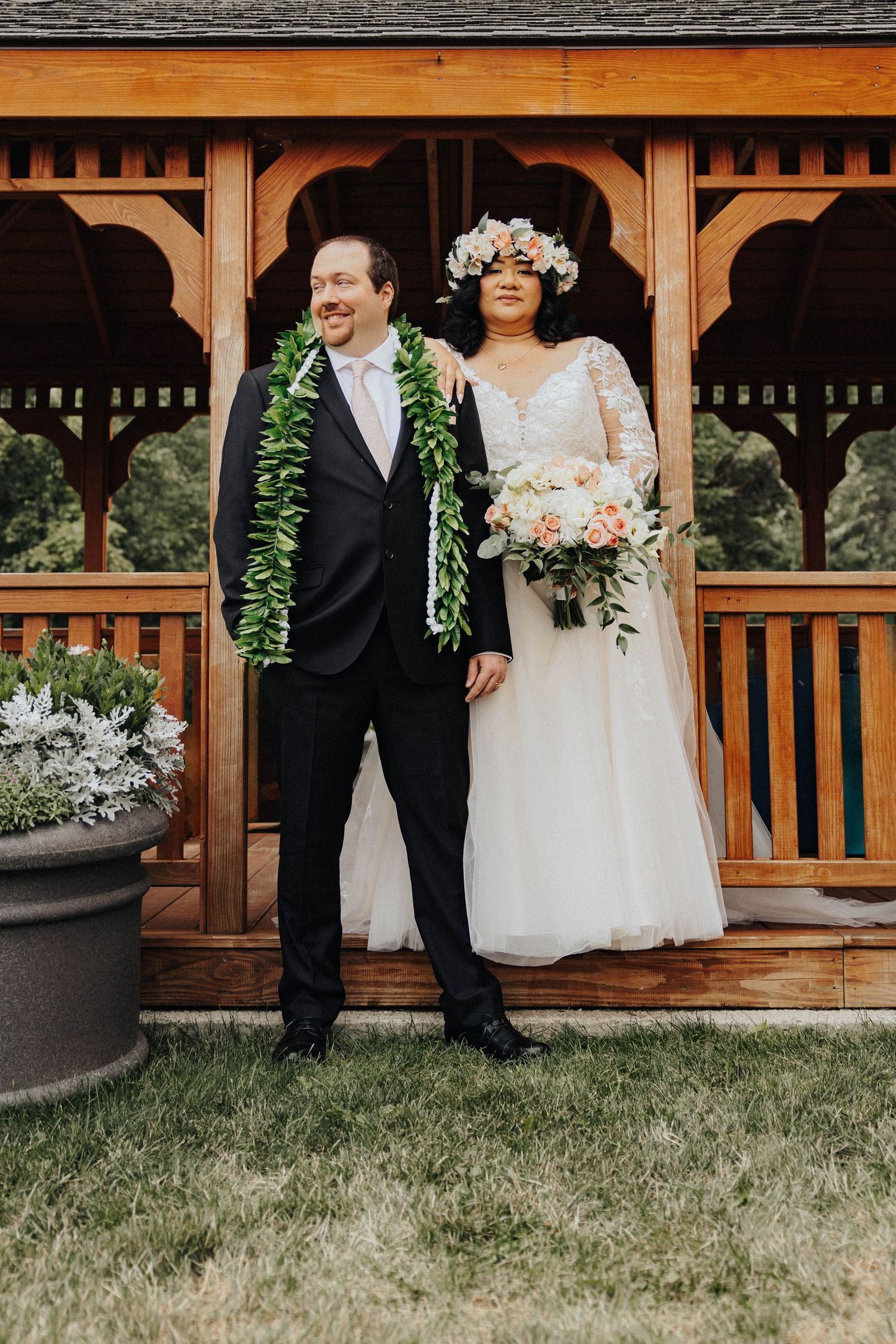A couple in wedding attire poses on a wooden gazebo with traditional Hawaiian leis and floral decorations.