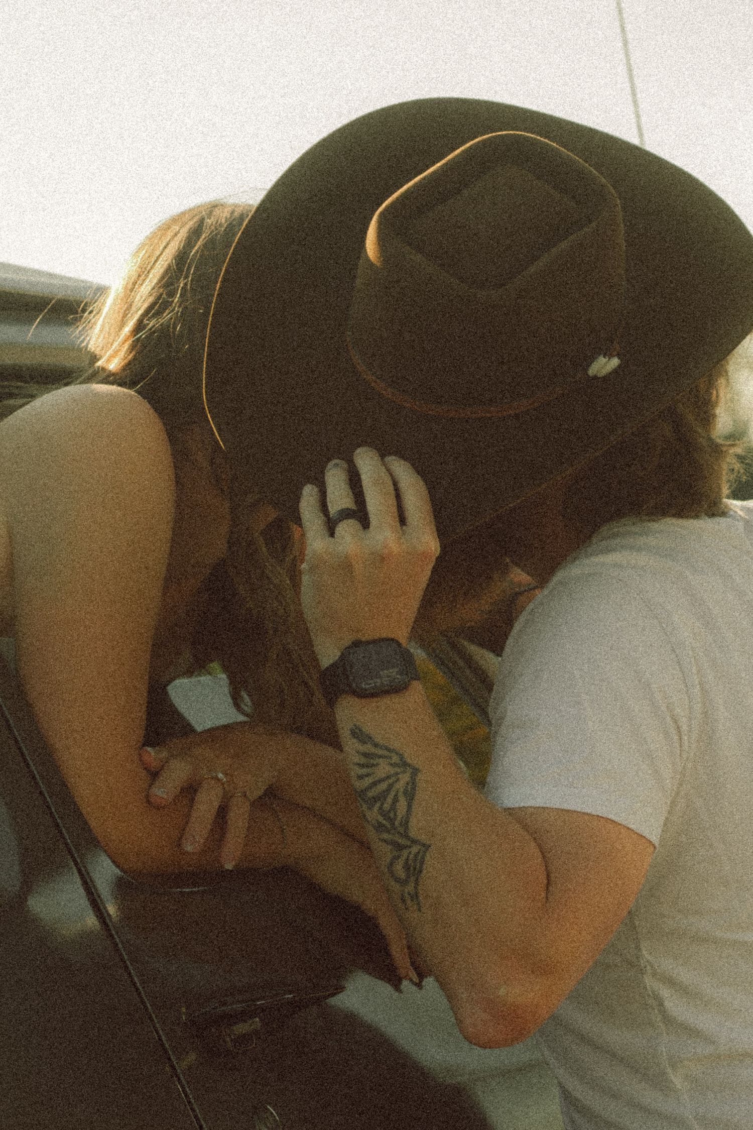 Couple sharing a kiss, man wearing black cowboy hat and watch with butterfly tattoo visible on arm.