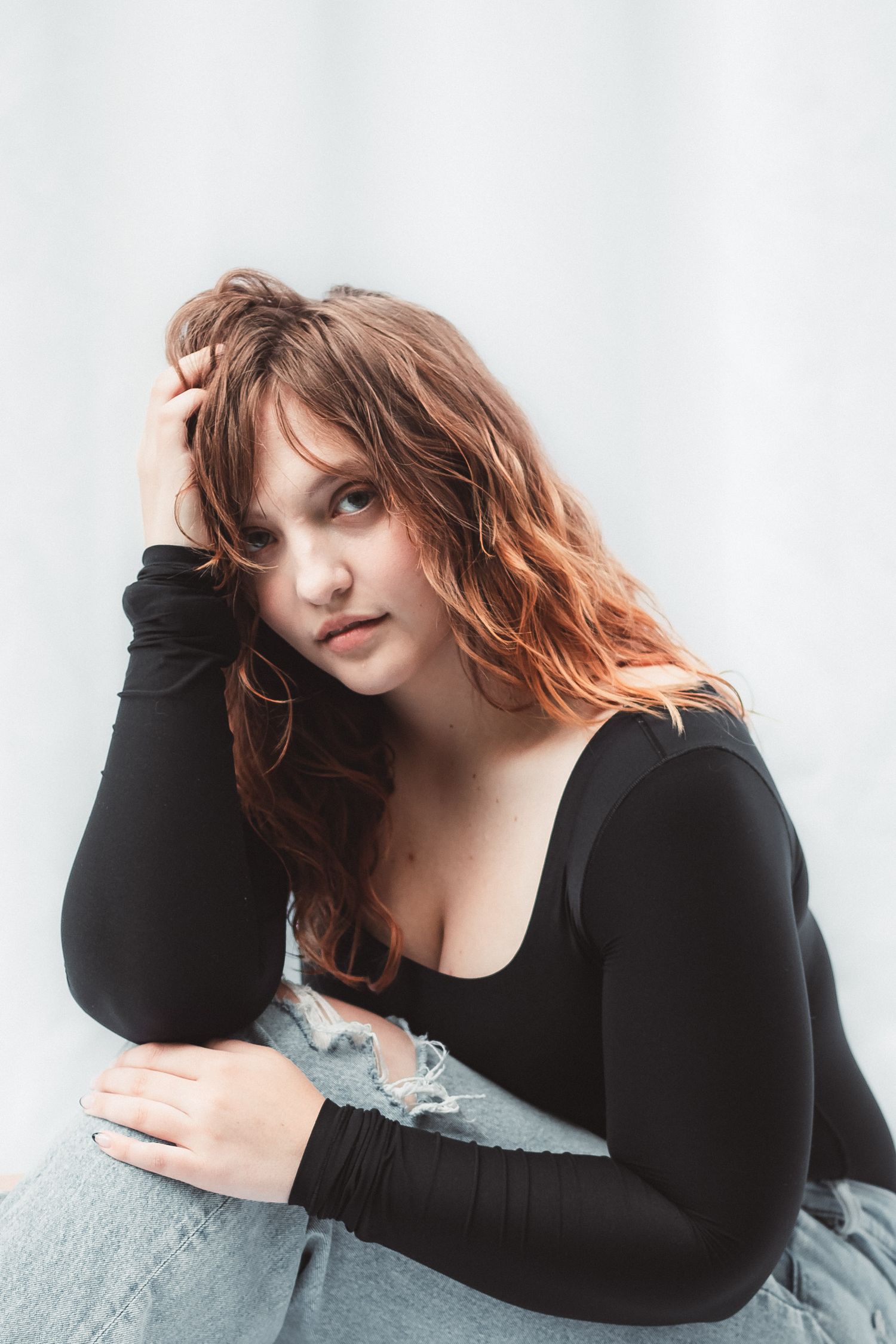 Young woman with wavy auburn hair in black top and ripped jeans poses casually against a white background.