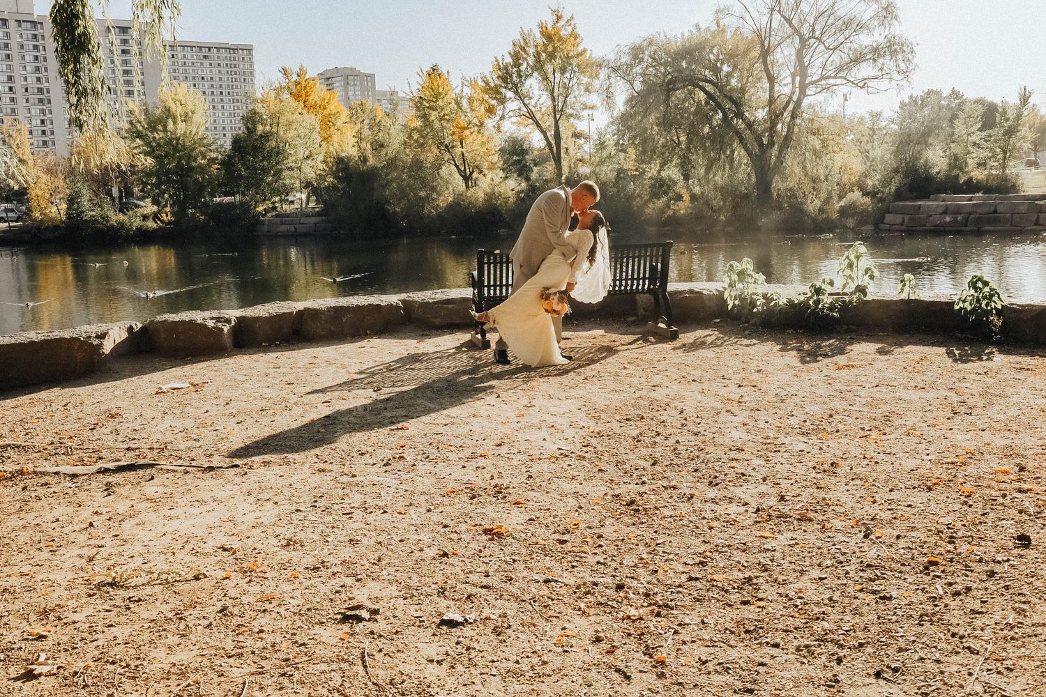 A couple poses together in an autumn park setting with golden trees and a lake in the background.