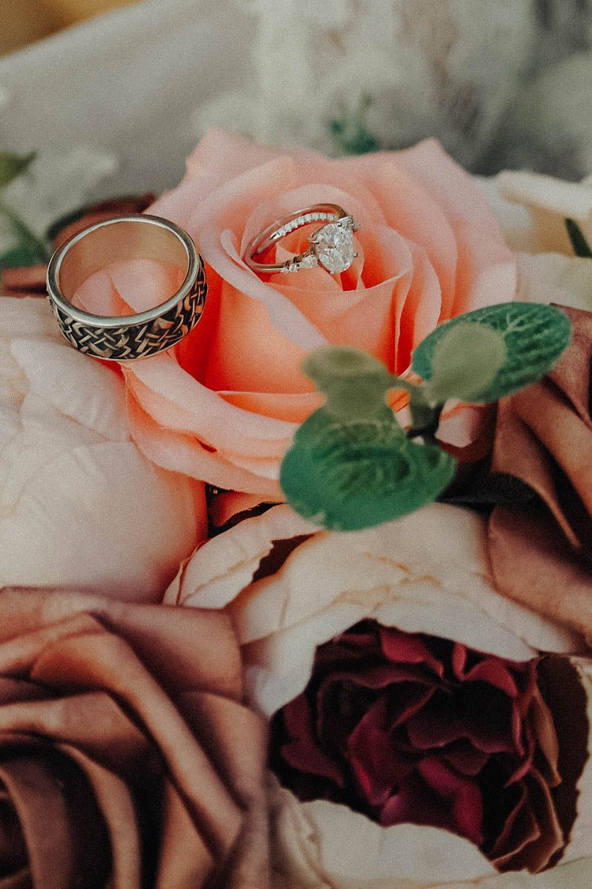 Wedding rings nestled in a peach-colored rose surrounded by burgundy roses and green foliage in romantic lighting.